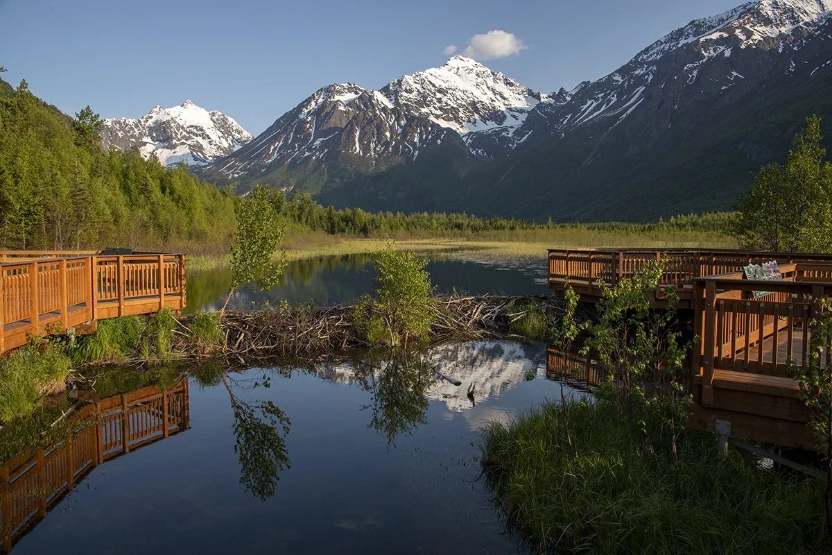 Wooden observation decks overlooking a calm lake with snow-capped mountains and green trees in the background.