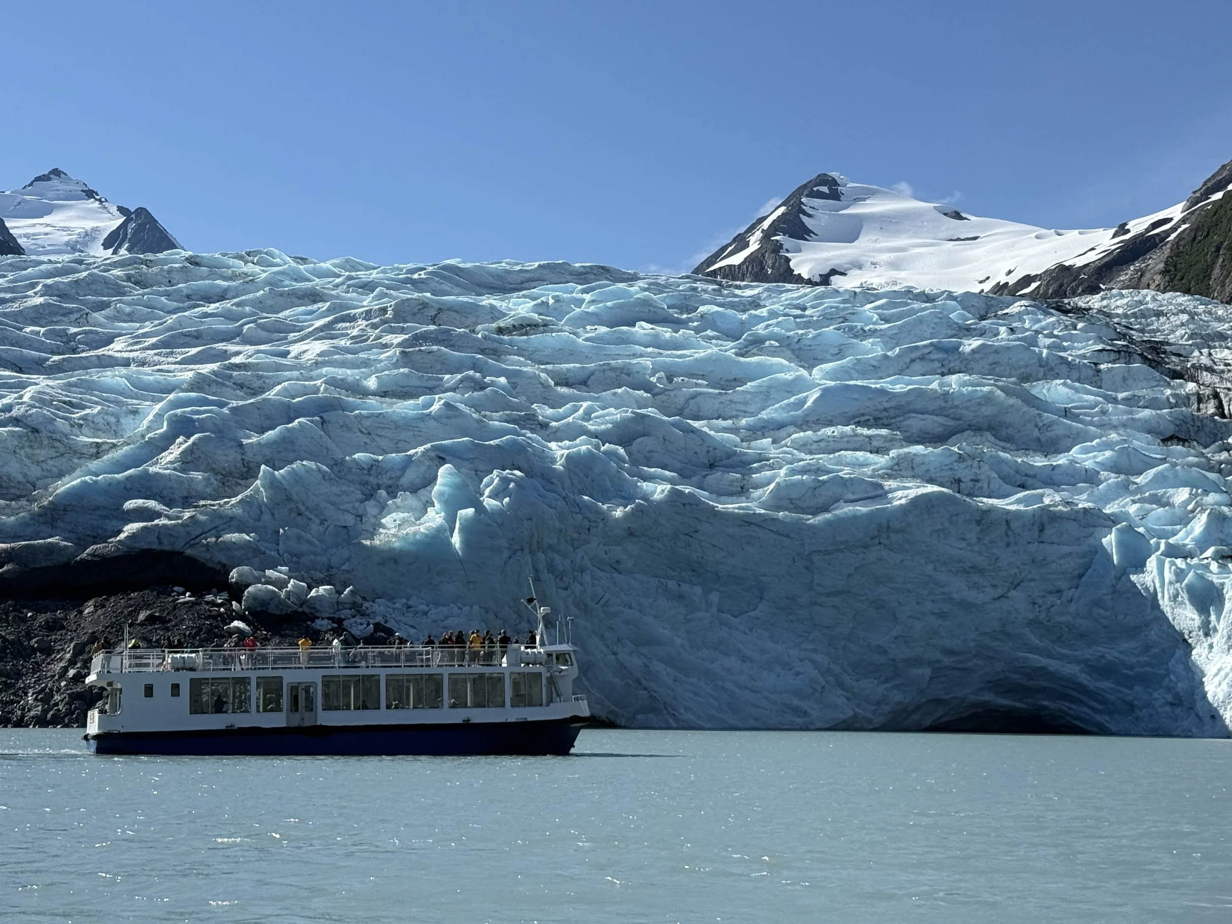 A white boat with large windows and a group of tourists on its upper deck floating on icy turquoise water, with a glacier and snow-covered mountains under a clear blue sky in the background.