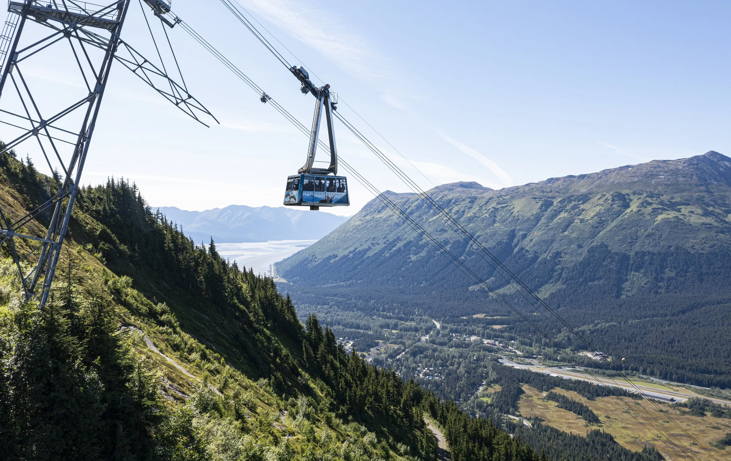 A cable car traveling across a mountain landscape with lush green hills and distant mountains under a clear blue sky.