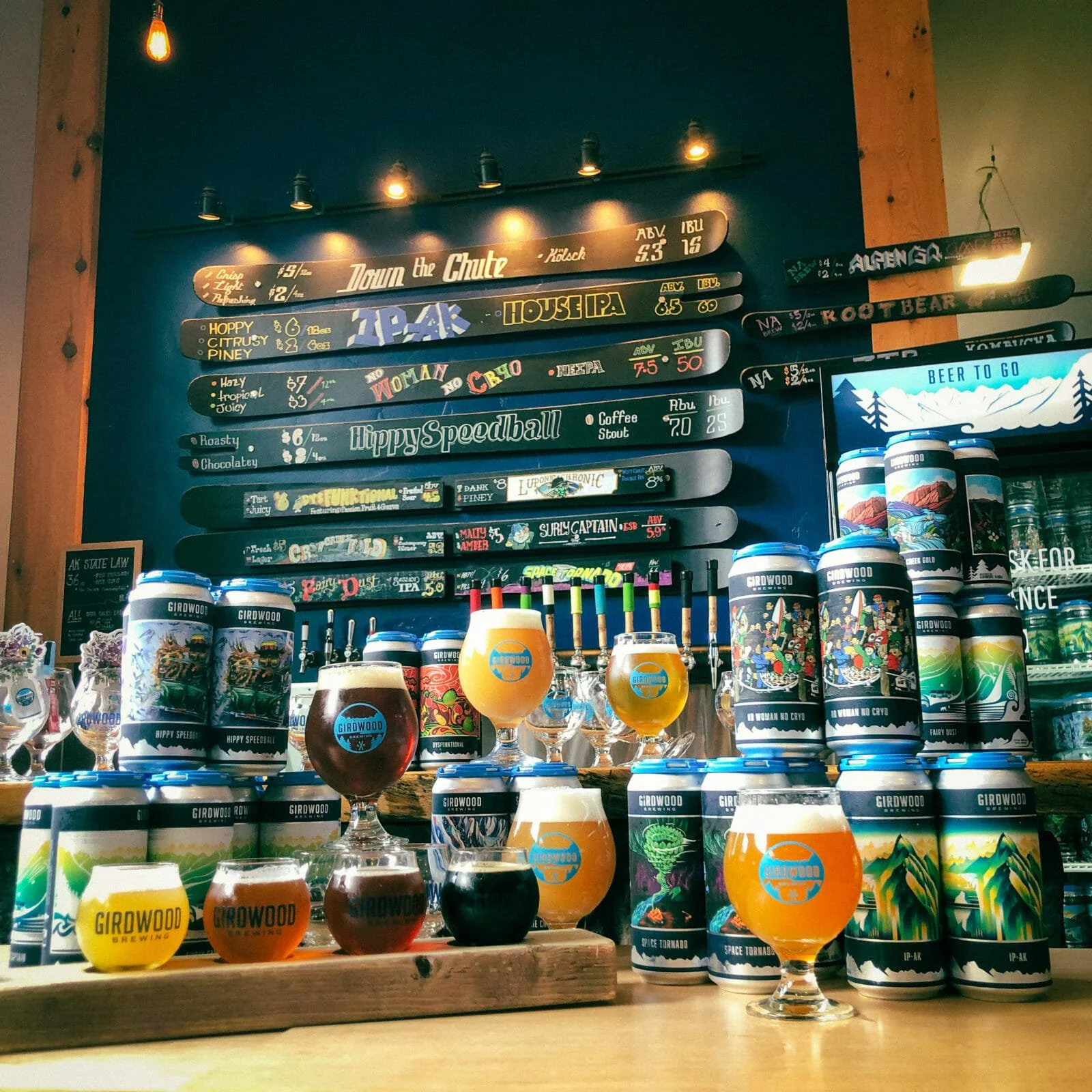 Various craft beers in colorful glasses and cans on a bar counter at a brewery, with a menu board listing different beer options in the background.