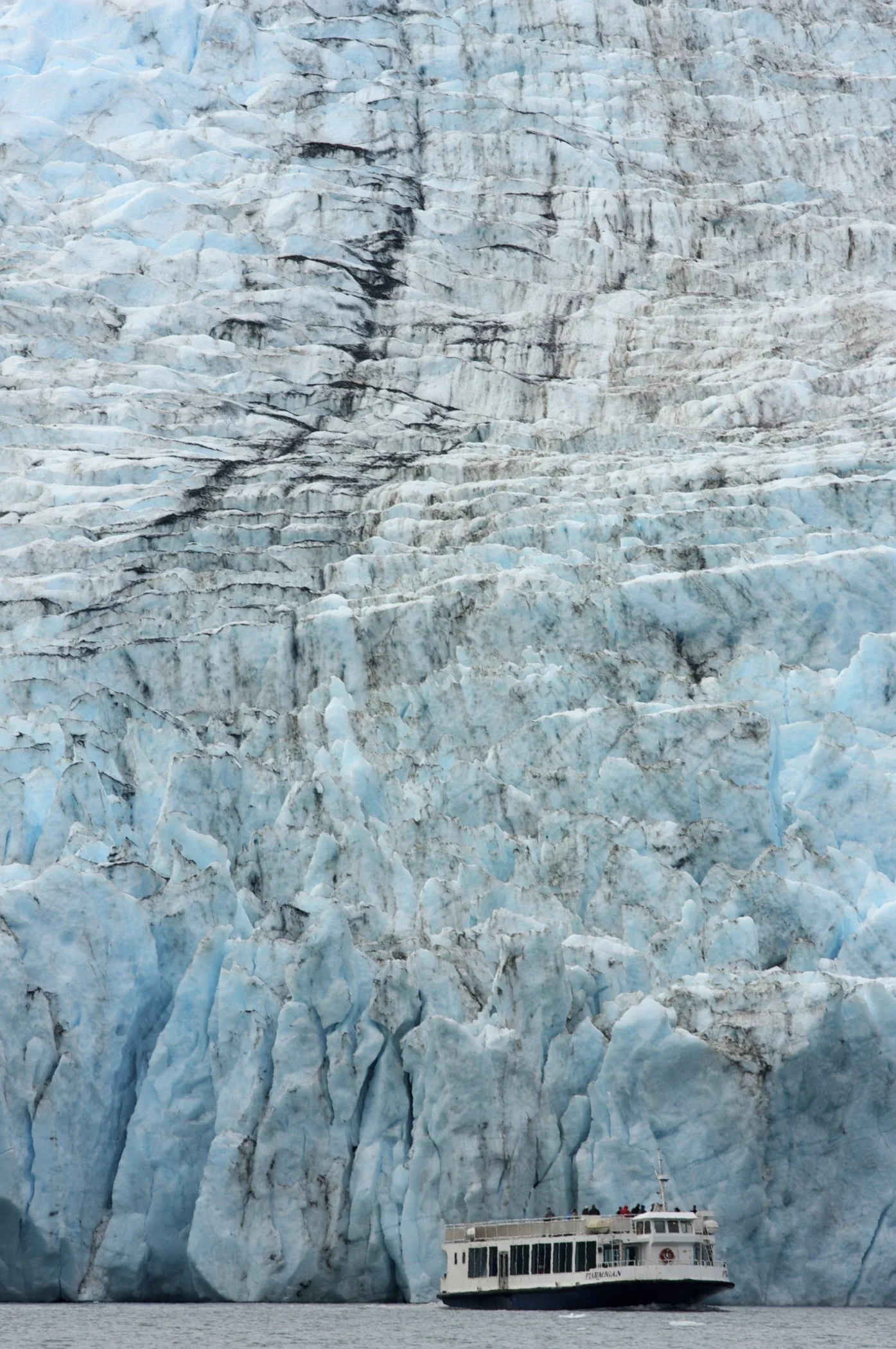 A boat approaching a large glacier with blue and white ice and dark streaks of dirt or volcanic ash on its surface.