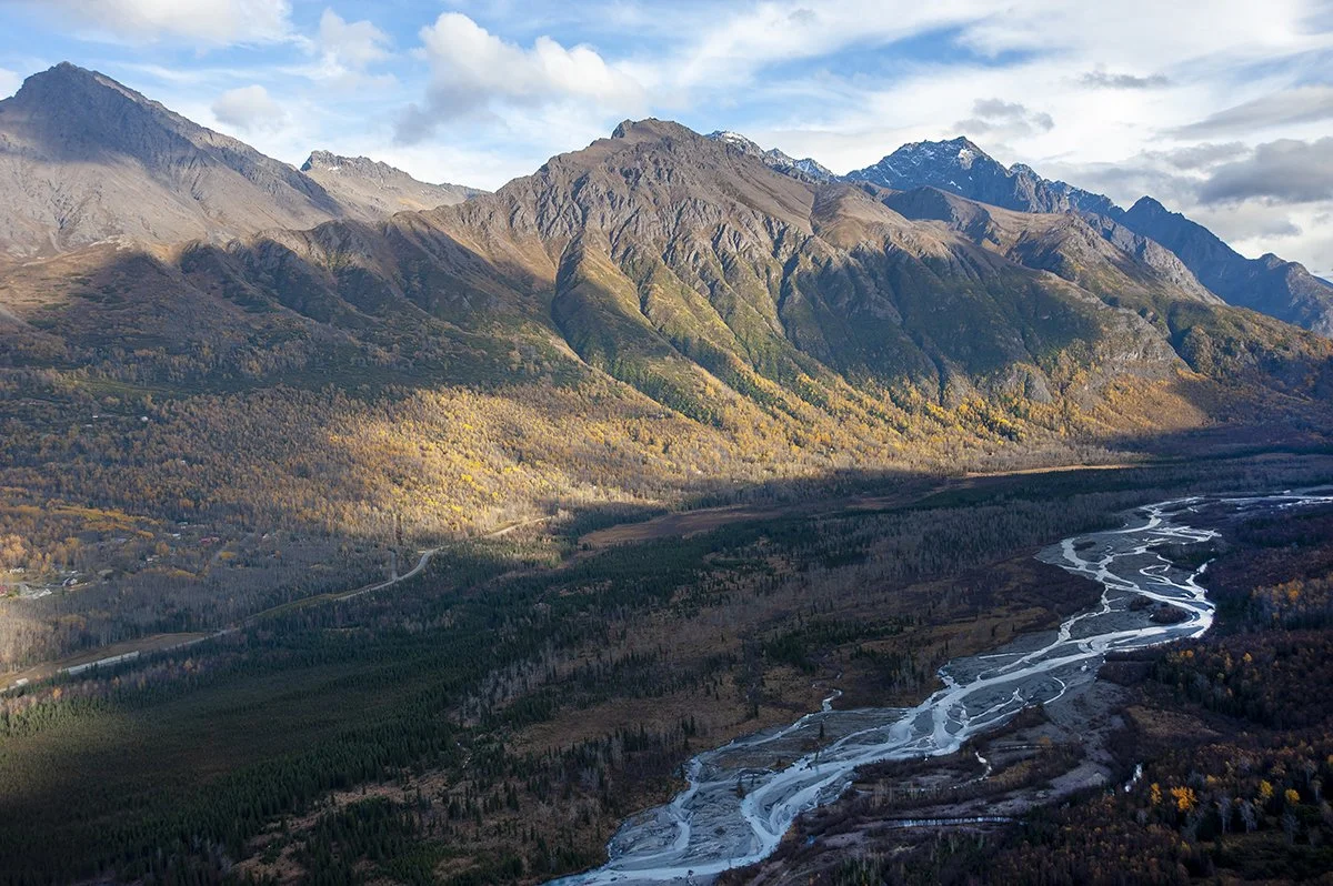 Aerial view of mountains with snow patches, forested slopes, and a winding river in the valley during daytime.