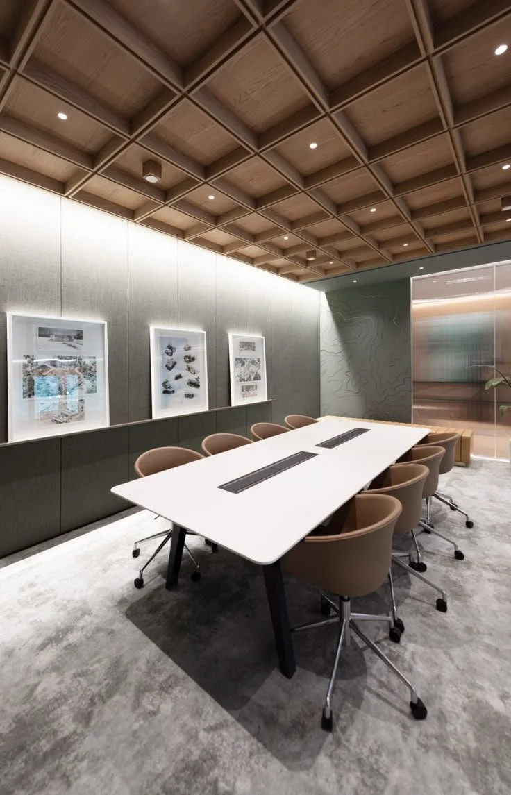 Modern conference room with a white table, beige rolling chairs, gray walls, wooden ceiling, and framed artwork.