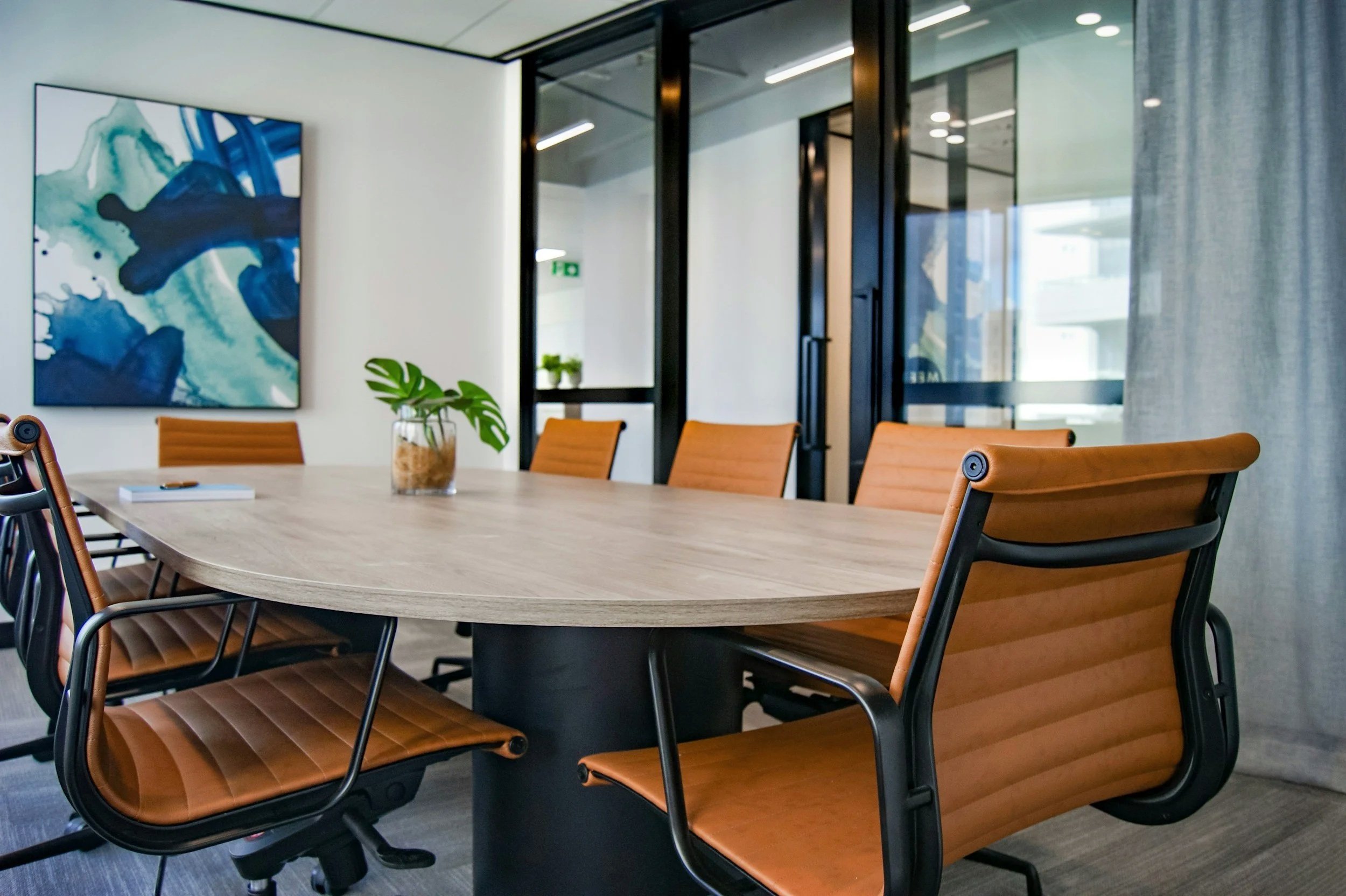 Modern conference room with a wooden table, tan leather chairs, a glass of plant, abstract artwork, and glass walls with curtains.