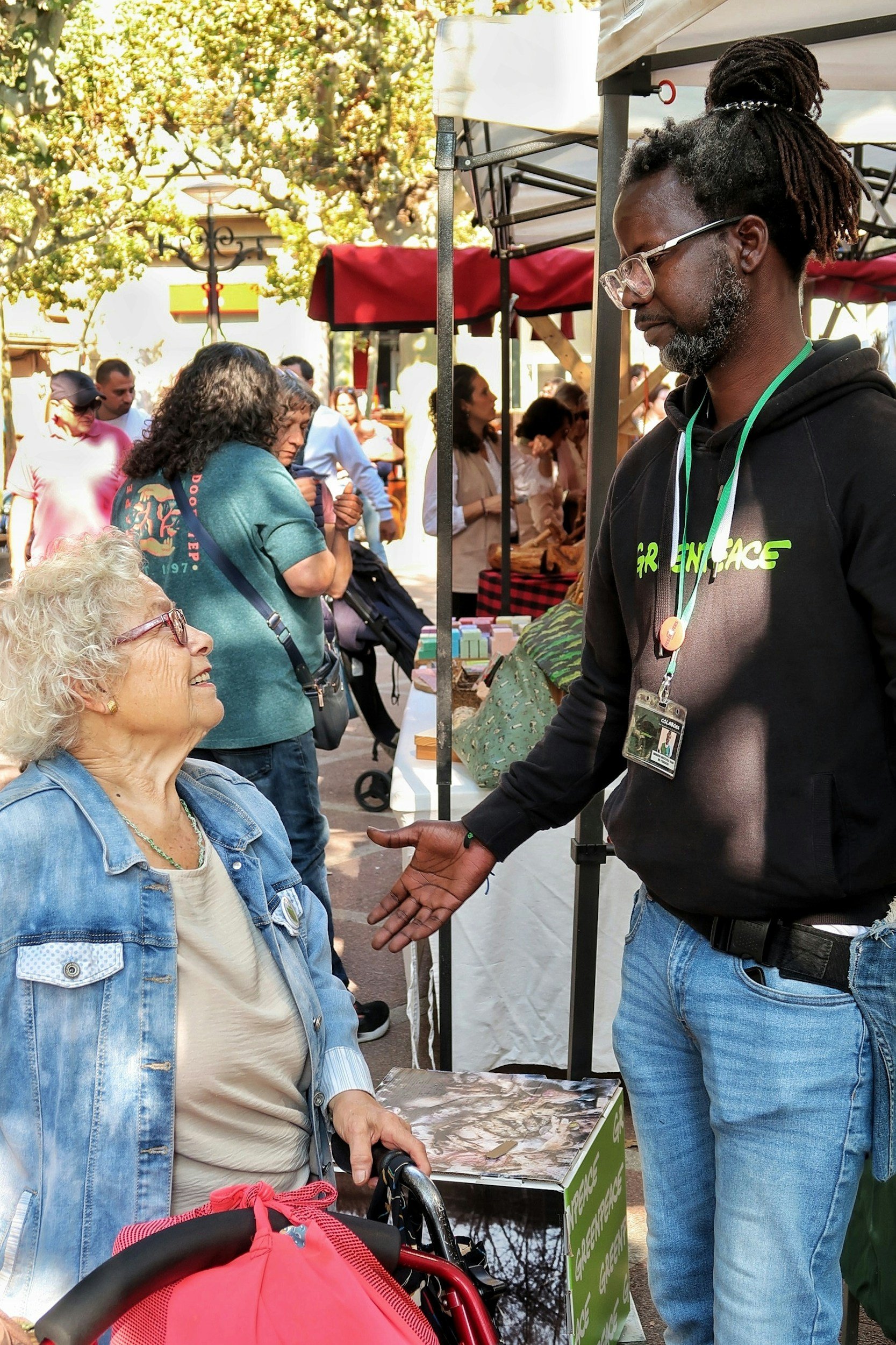 A man with glasses and dreadlocks is talking to an elderly woman in a denim jacket at an outdoor market or fair. The scene includes multiple people, tents, and trees in the background, with sunlight filtering through the foliage.