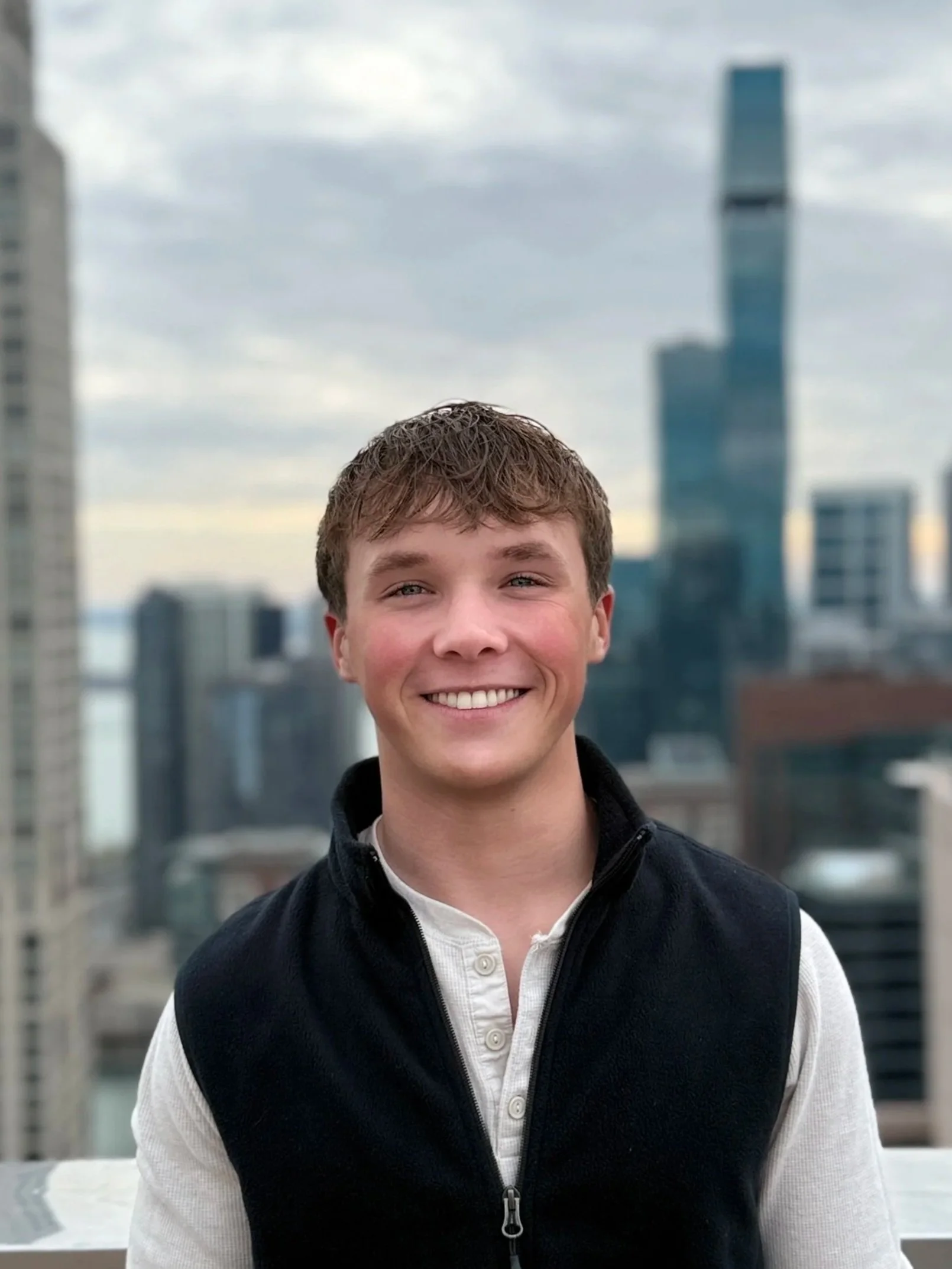 A young man smiling outdoors on a rooftop with a cityscape in the background, including tall buildings and a cloudy sky.
