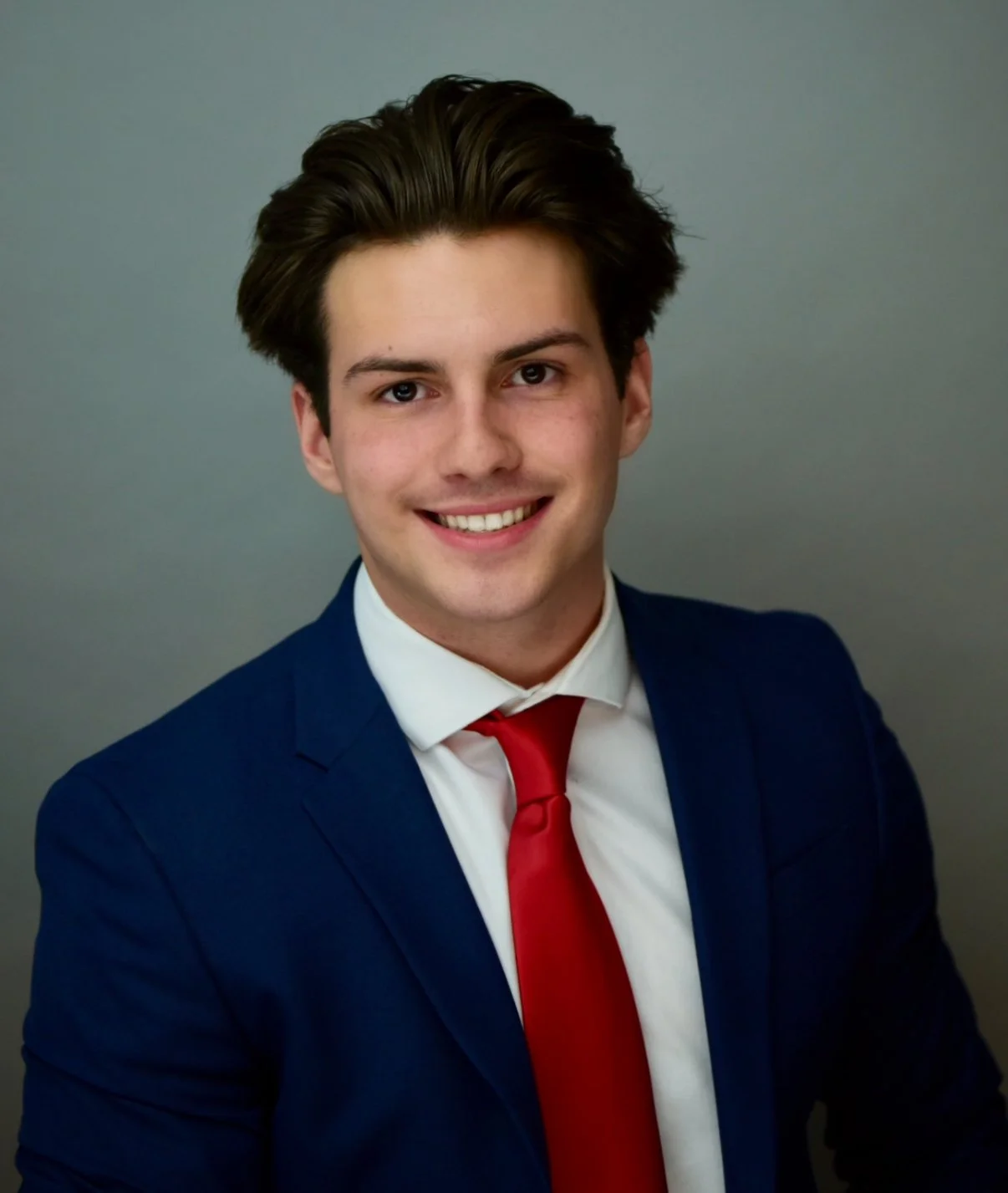 Young man in a navy suit, white shirt, and red tie smiling against a plain gray background.