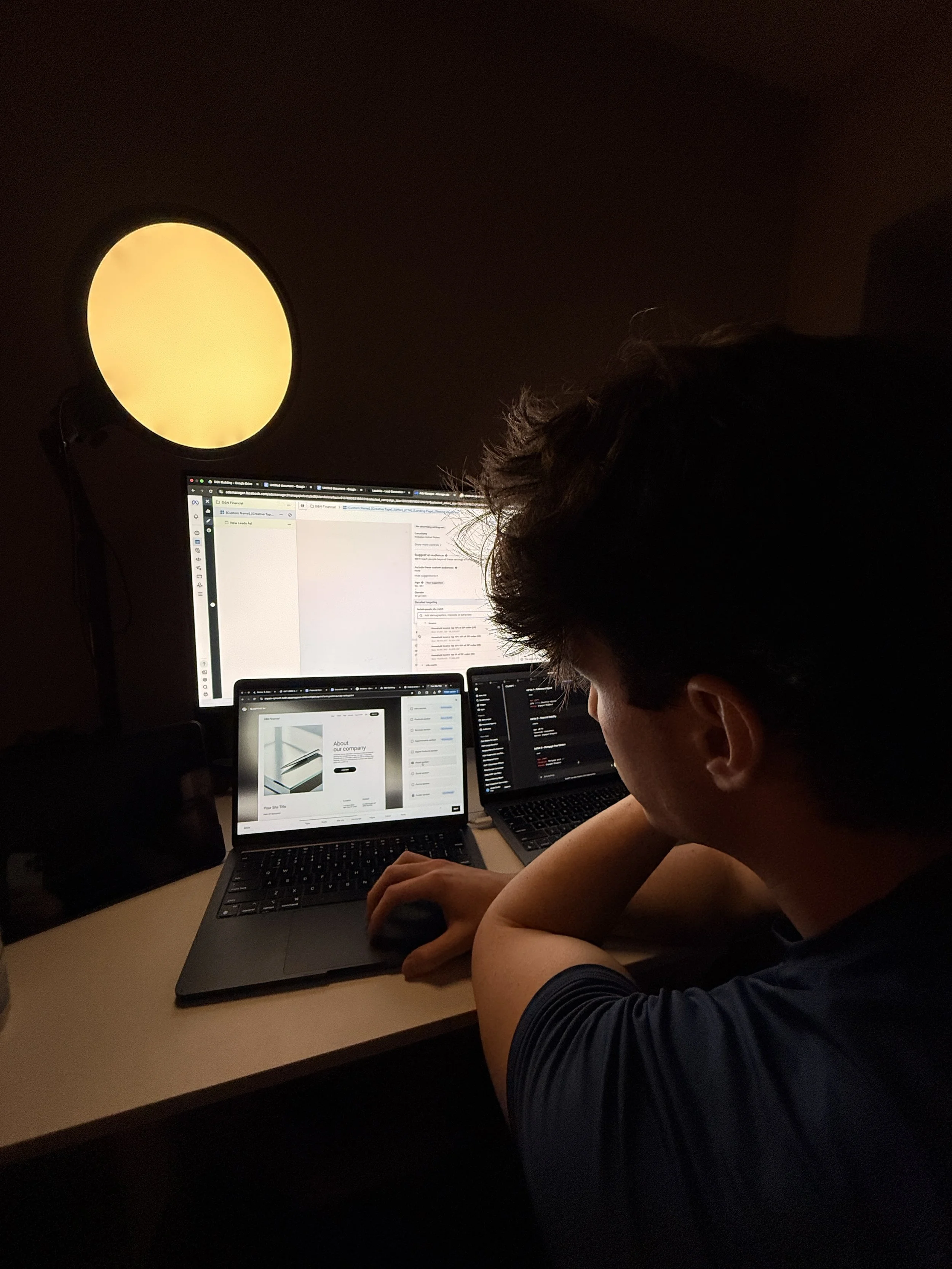 A person working at a desk with multiple laptop screens, in a dimly lit room, illuminated by a large round lamp on the wall.
