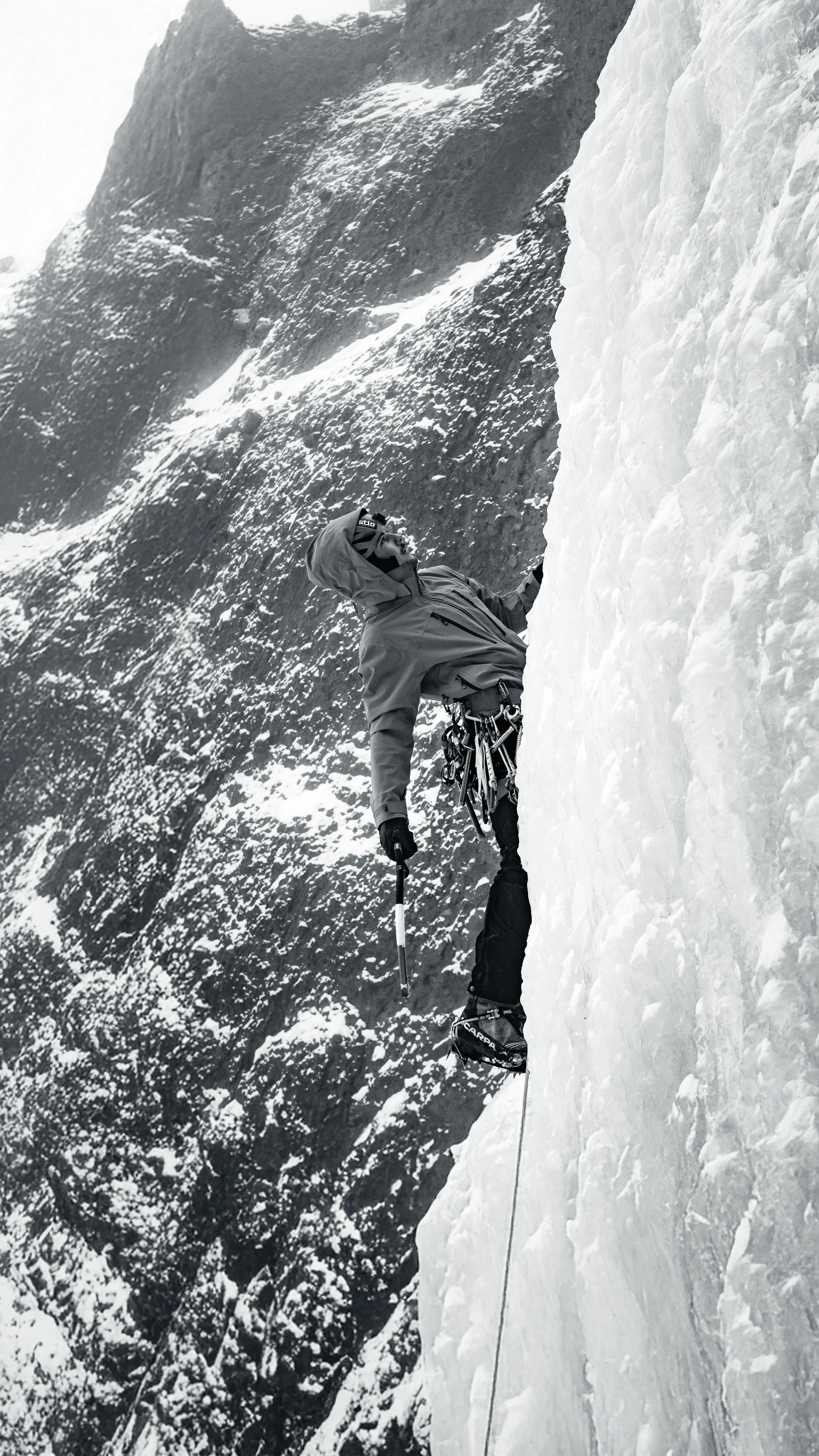 Person ice climbing on a steep frozen wall, wearing a hooded jacket and harness, with a mountain rugged background.