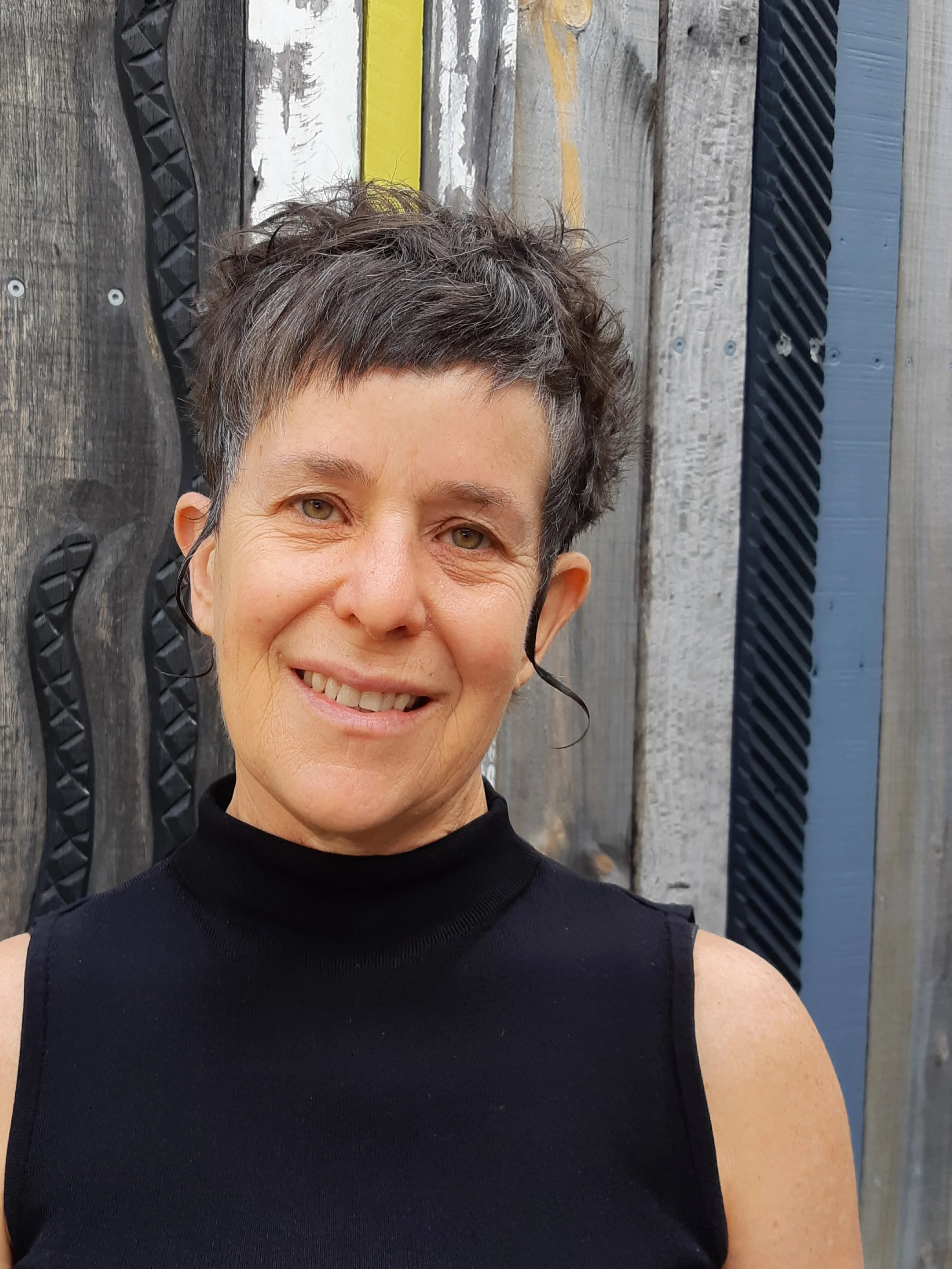 A woman with short, dark curly hair smiling at the camera, standing outdoors in front of a weathered wooden fence with black decorative elements.