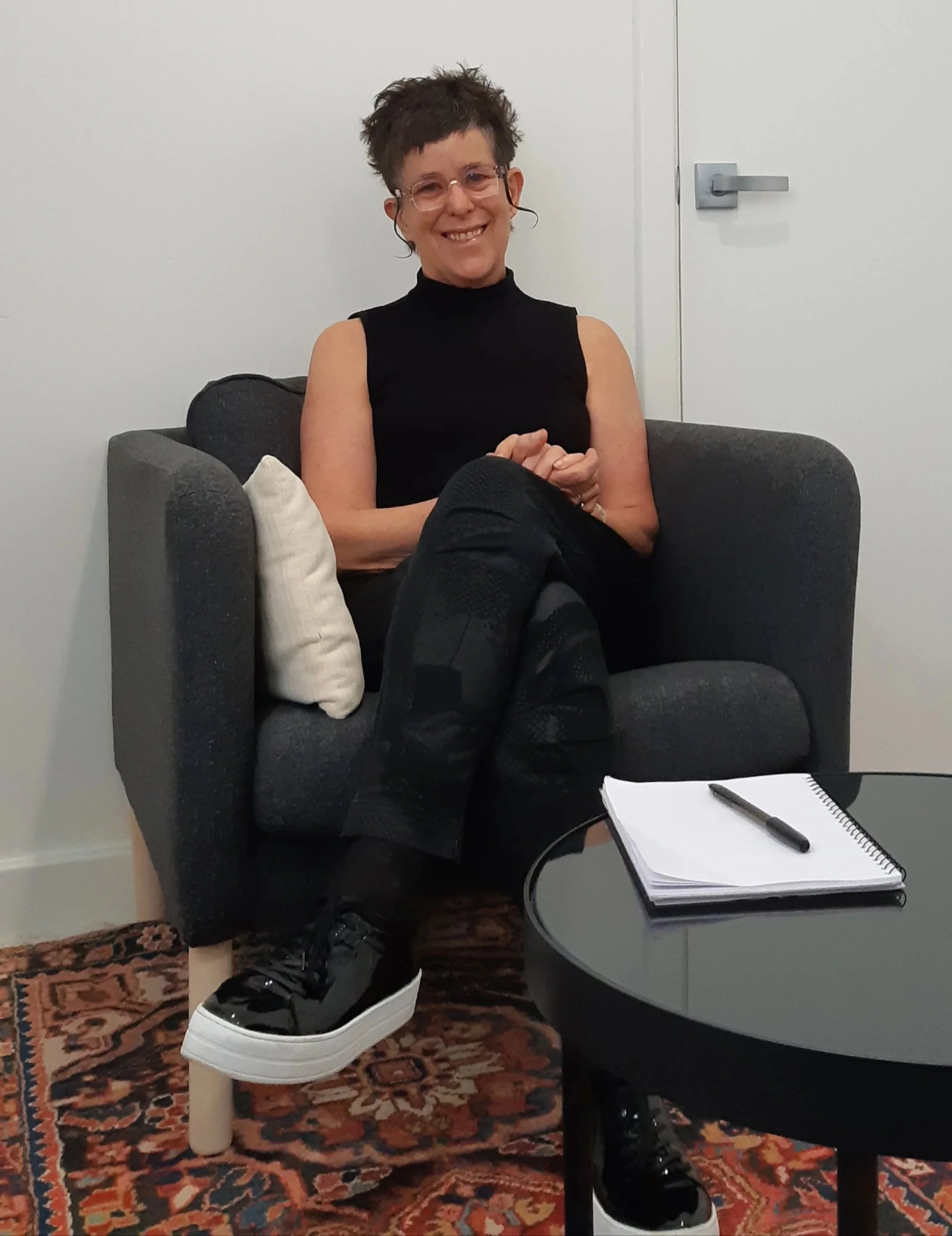 A woman with short, spiky hair wearing glasses, a black sleeveless top, patterned black pants, and black sneakers with white soles, sitting on a gray armchair with a white pillow, smiling at the camera. There is a notebook and pen on a round black table in front of her.