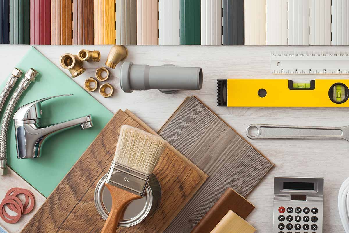 A collection of home improvement and painting tools on a white surface, including paint swatch samples, a paintbrush on a can, a faucet, brass fittings, a level, a ruler, and a calculator.