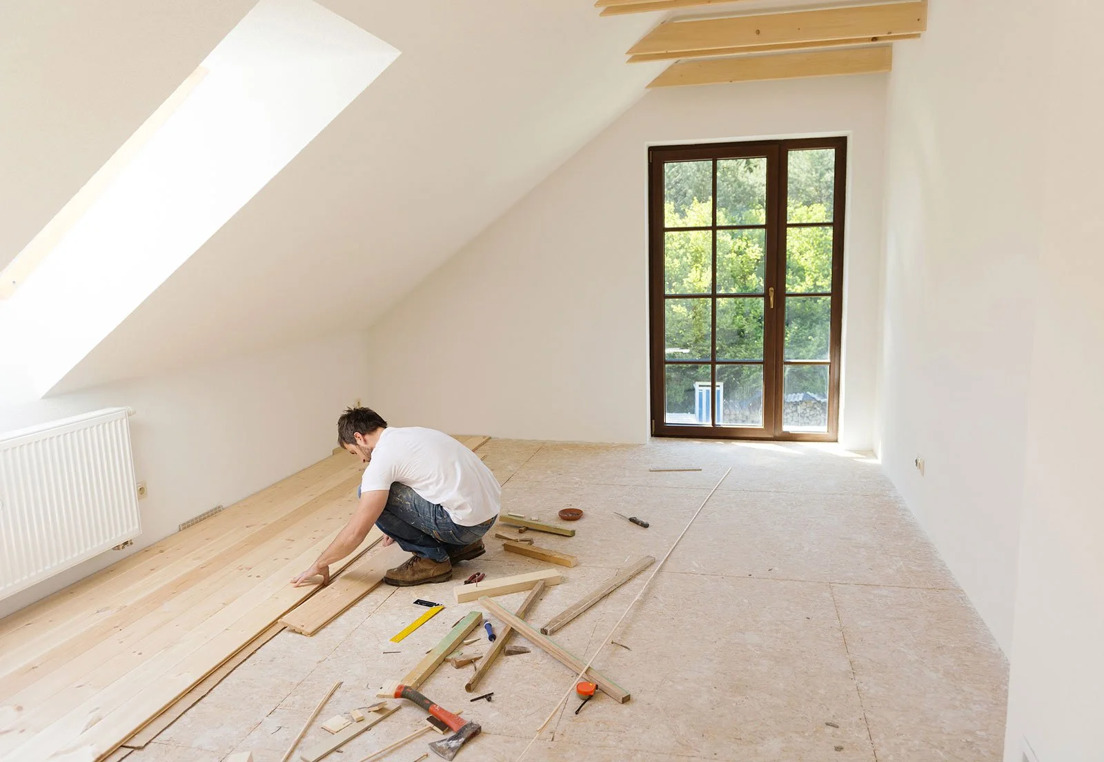 A man working on installing wooden flooring in an attic room with a sloped ceiling and a large window showing green trees outside.