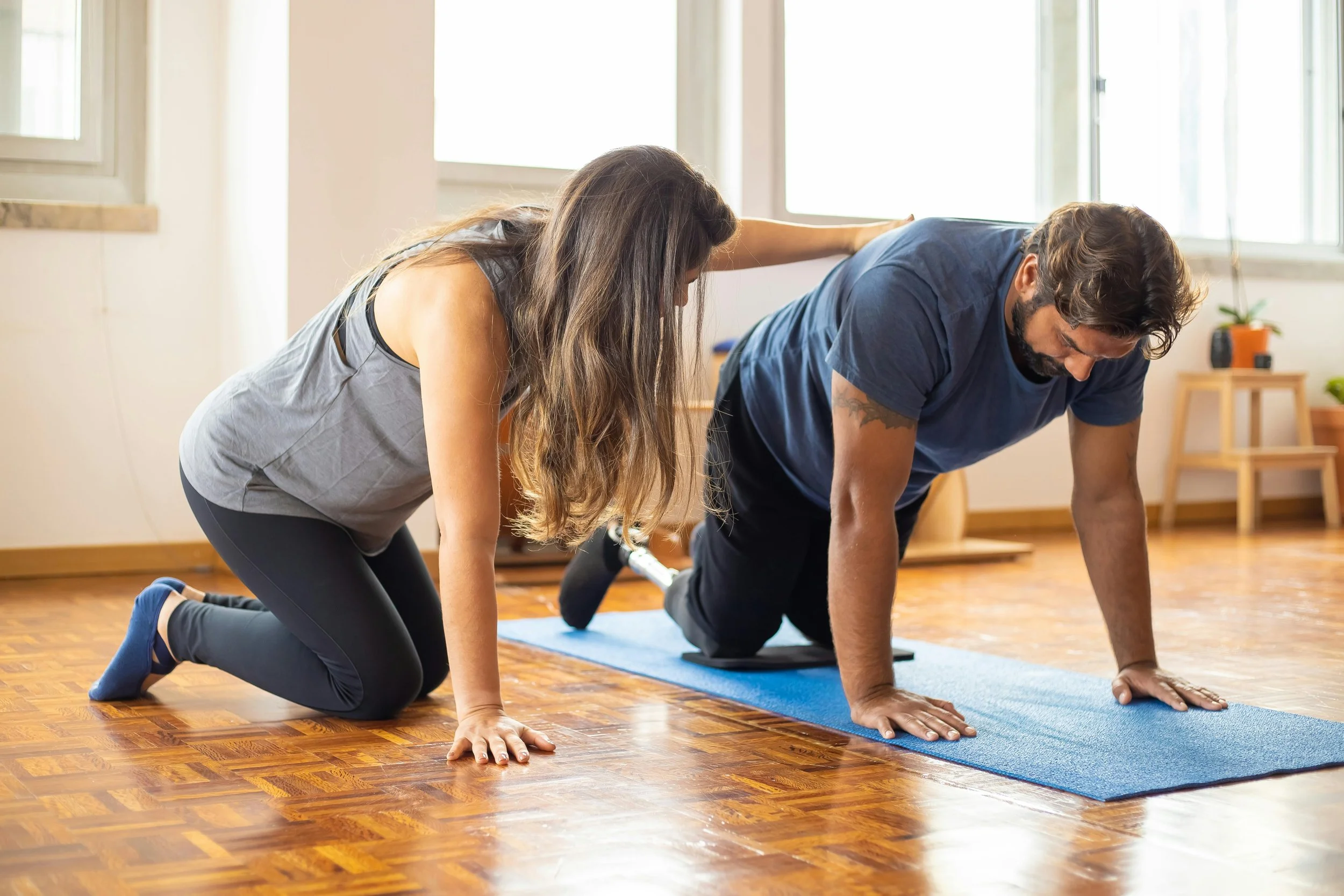 A woman and a man doing a plank exercise together on a yoga mat in a bright room.