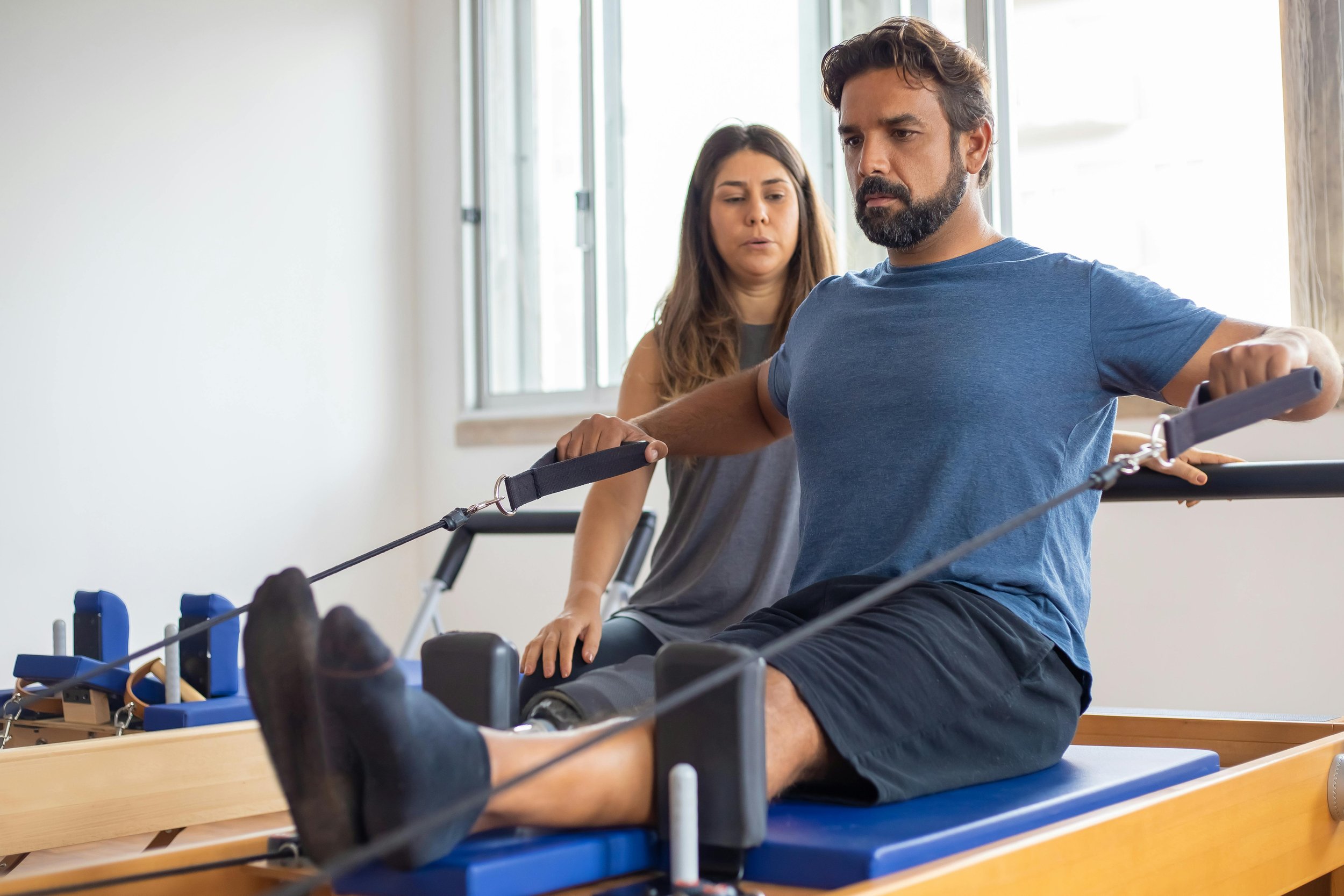 A man is doing physical therapy exercises on a reformer machine, with a woman assisting him in a therapy room filled with natural light from large windows.