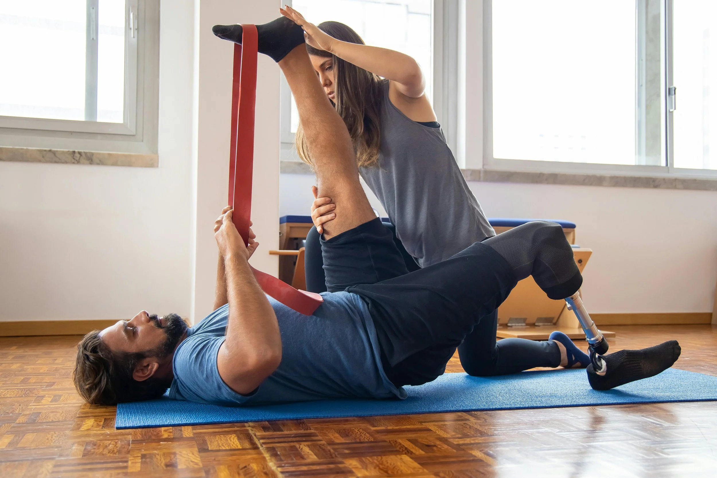 A woman assists a man with a prosthetic leg during physical therapy on a blue exercise mat inside a room with large windows. The man is lying on his back and lifting his leg with a red resistance band while the woman supports him.