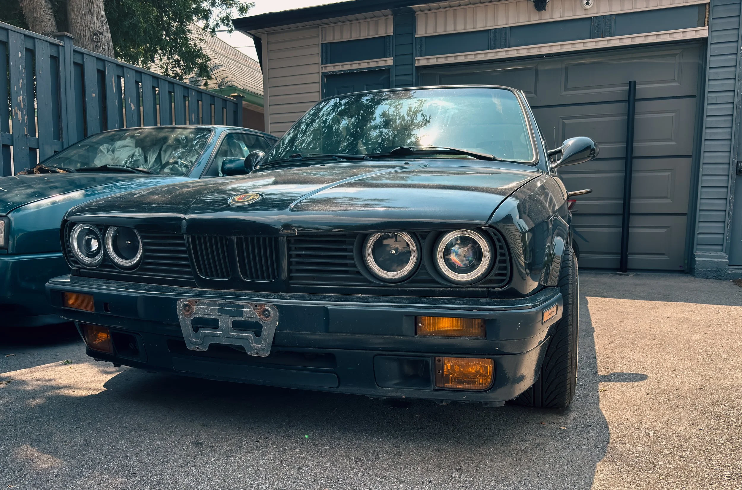 Black vintage BMW Z1 sports car parked in driveway next to a blue classic car, garage and wooden fence in background.