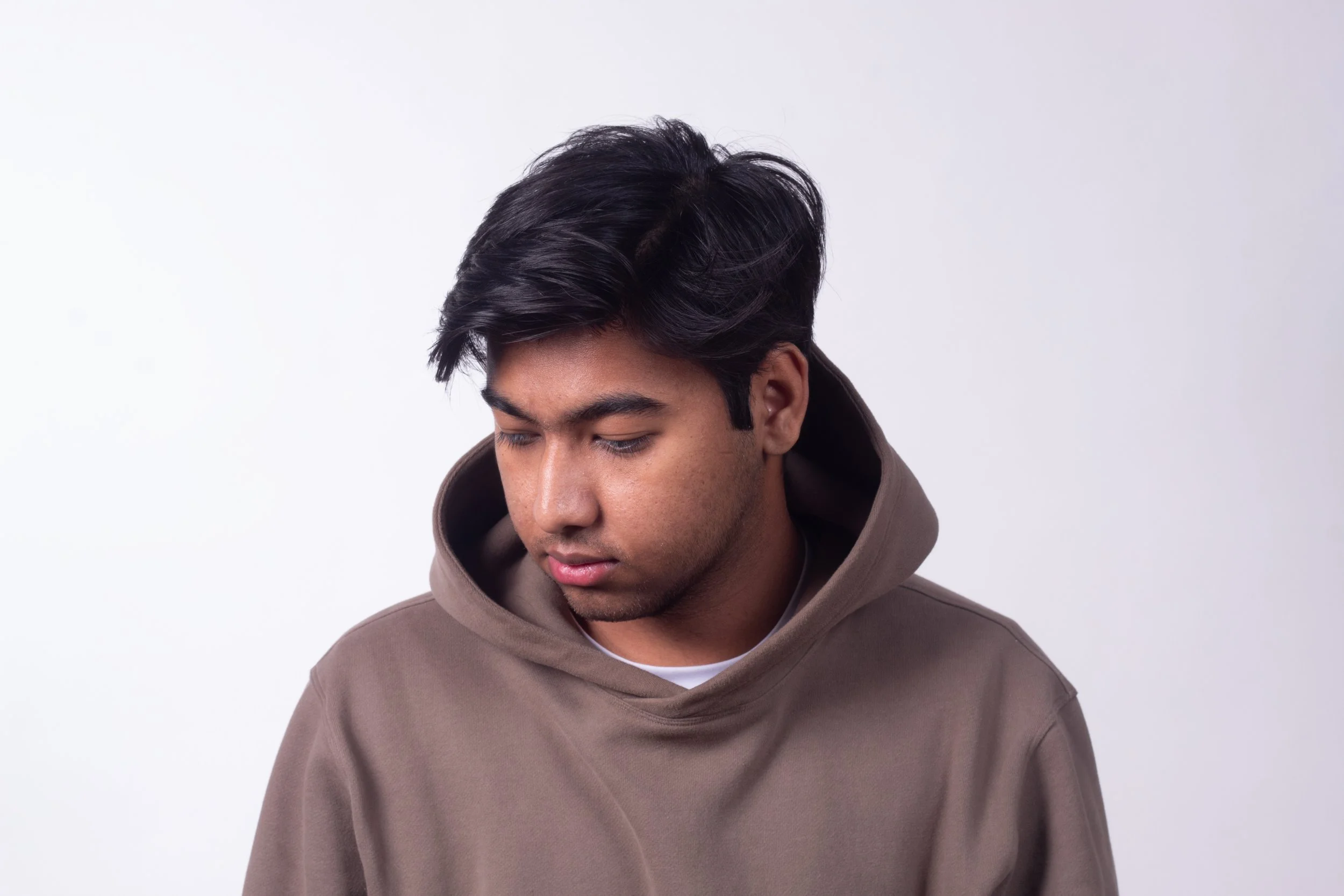 A young man with dark hair is looking down, wearing a brown hoodie, against a plain white background.