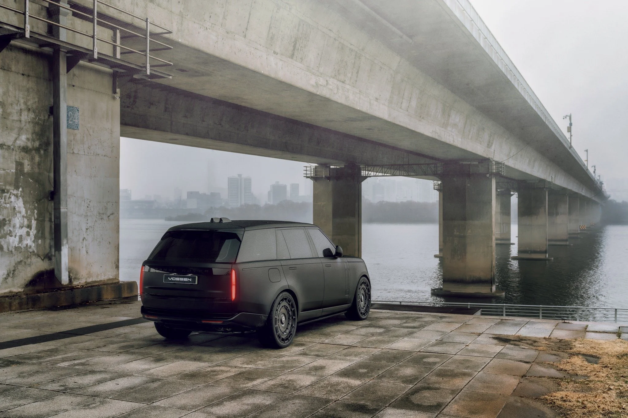 A matte black luxury SUV parked under a large bridge over a body of water, with a city skyline in the distance.