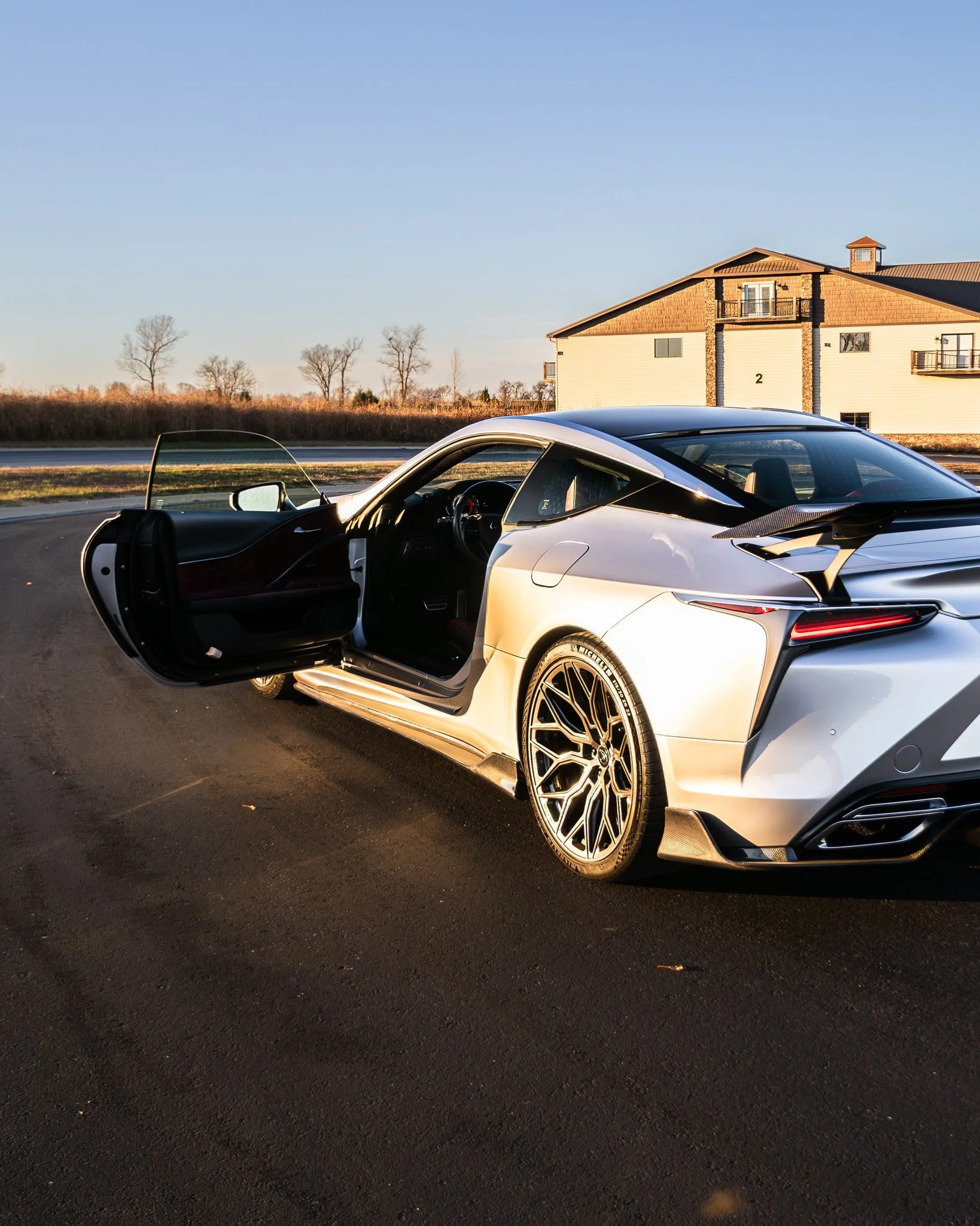 A silver sports car parked on an asphalt road with its driver's side door open, a modern house and leafless trees in the background, during late afternoon or early evening.