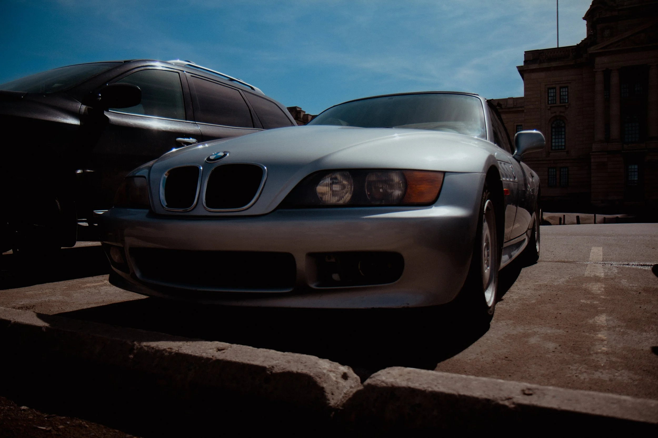 A silver BMW Z3 parked next to a black vehicle in a parking lot, with a historic brick building in the background under a blue sky.