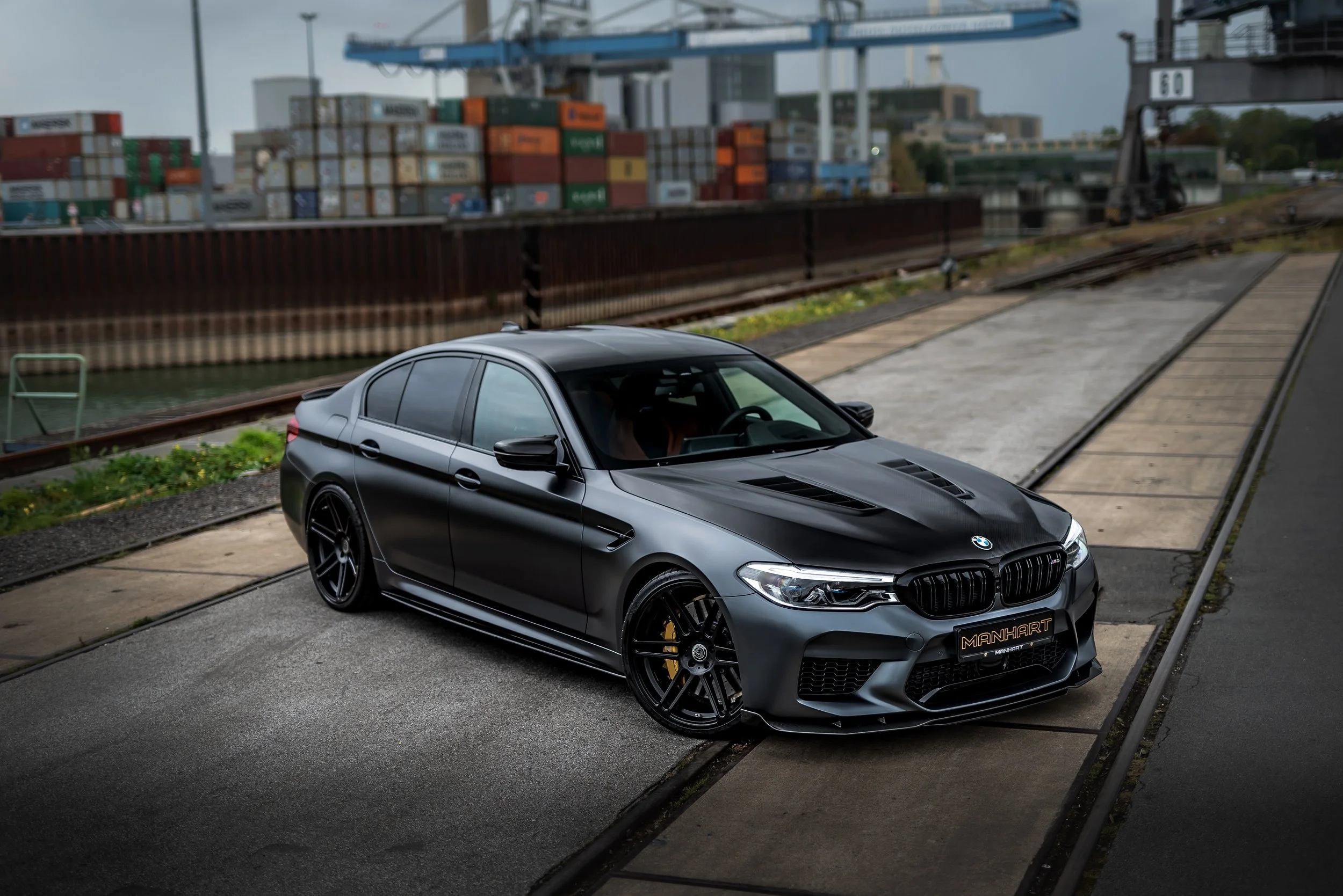 A black BMW sedan parked on a paved sidewalk near a shipping yard with stacked containers and cranes in the background.