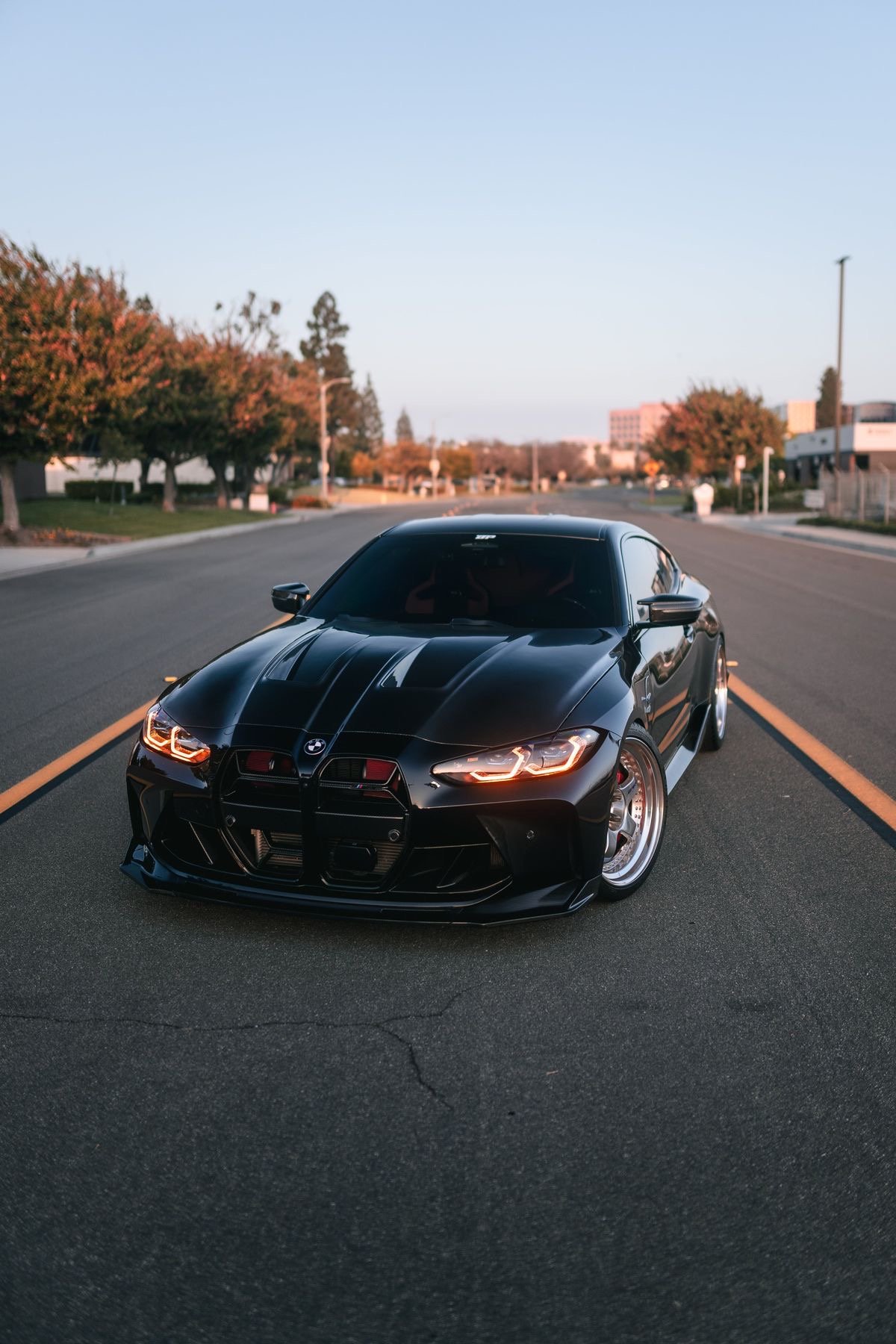 Black BMW M4 parked on an empty street during sunset with trees and buildings in the background.