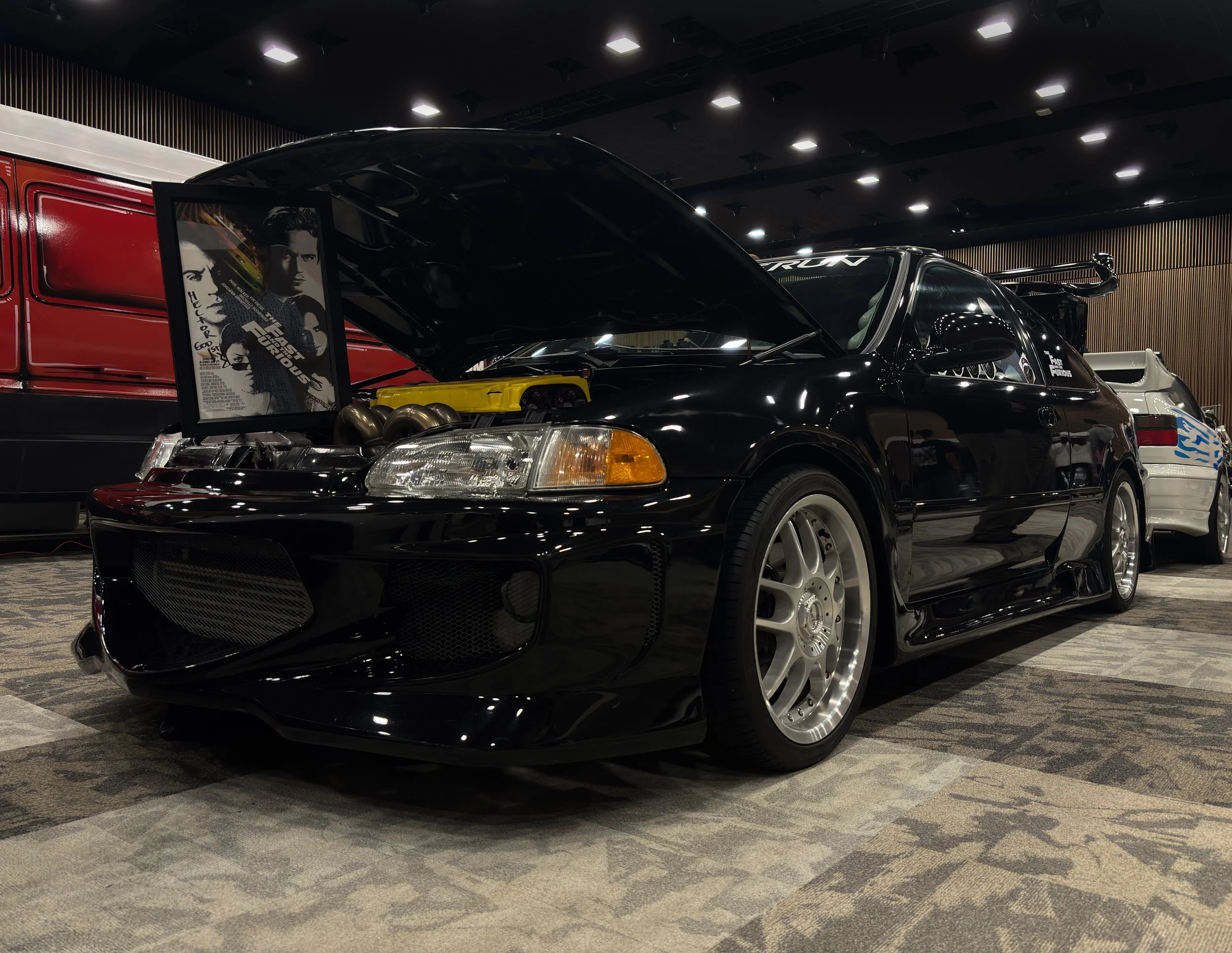 Black sports car with open hood on display in an indoor exhibit, with a poster for the movie Fast & Furious and other cars in the background.