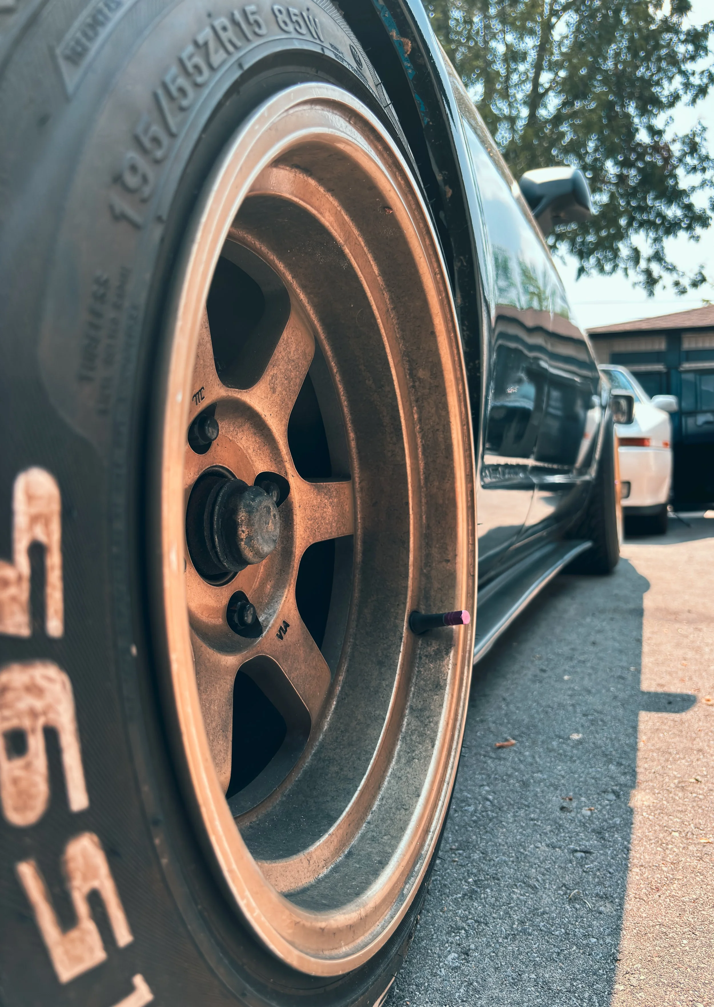 Close-up of a car tire with a brownish rim, parked on a driveway under a tree, with other cars and a house visible in the background.