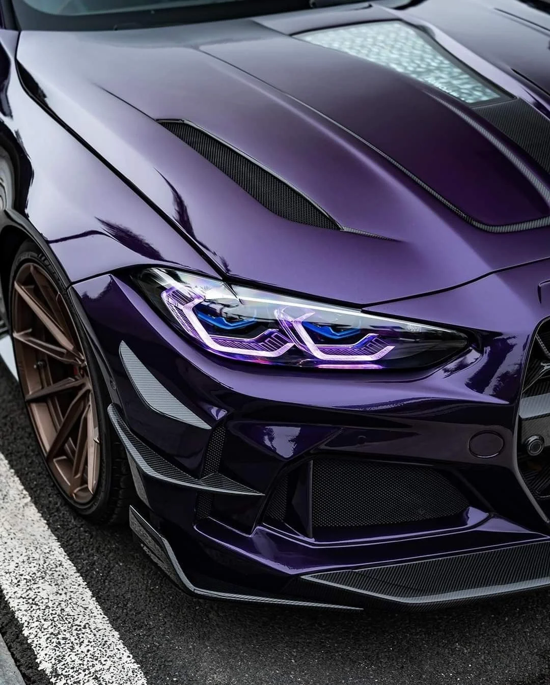 Close-up of a purple sports car with sleek design, carbon fiber accents, and illuminated headlights, parked on asphalt with white parking lines.