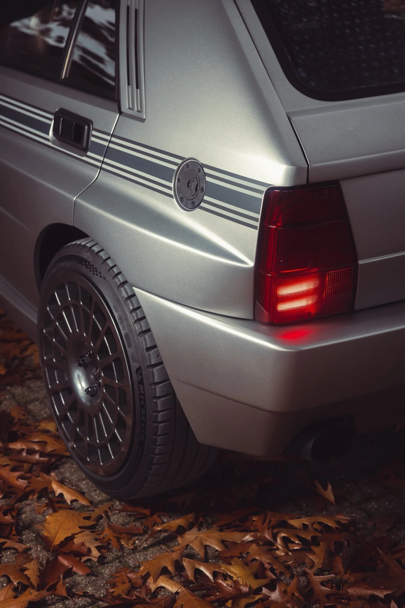 Close-up of the rear side of a silver car, showing the taillight, black wheel with intricate spokes, fuel cap, and some orange autumn leaves on the ground.
