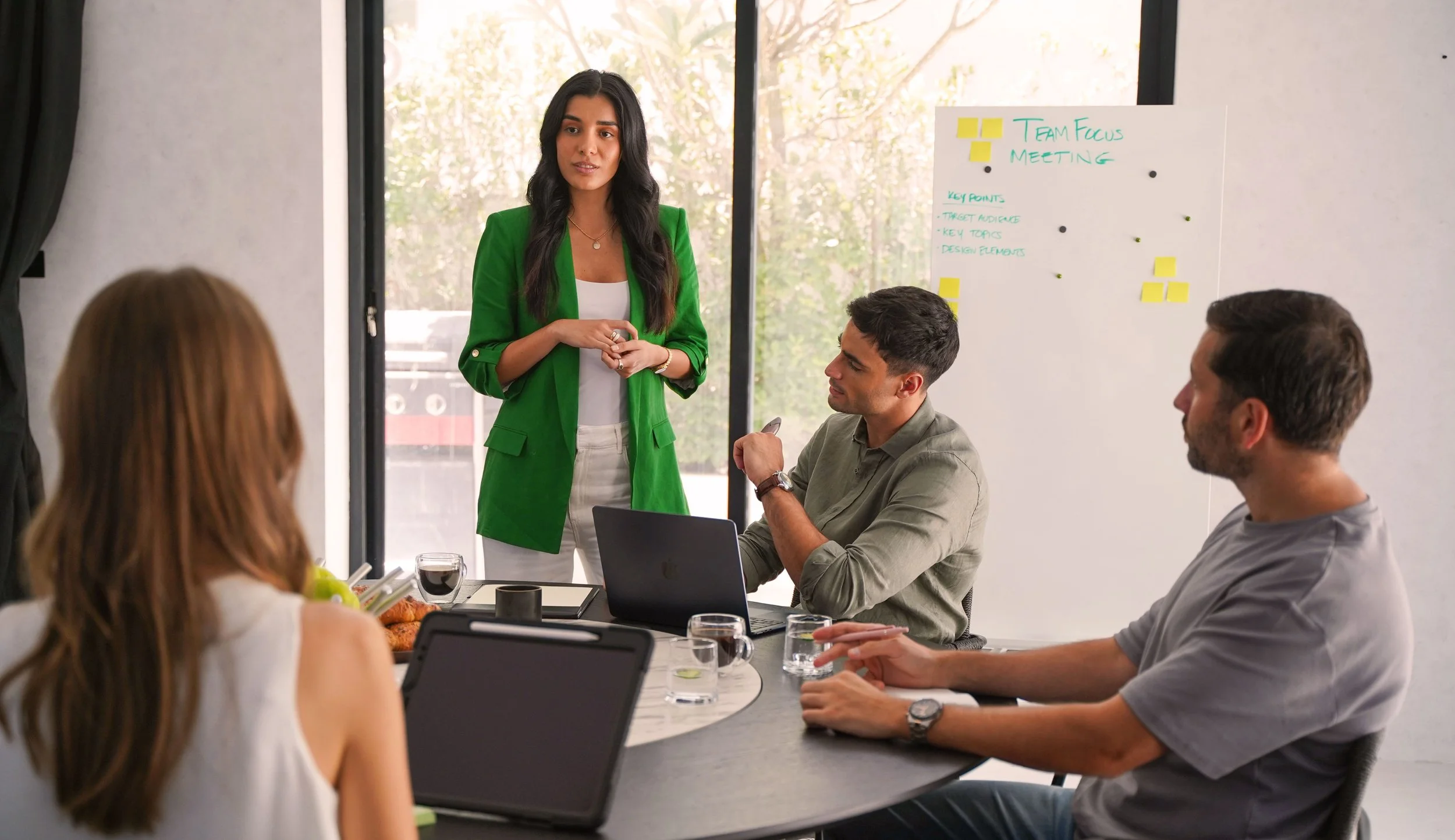 A woman in a green blazer giving a presentation to a group in a meeting room with a whiteboard that says 'Team Focus Meeting' and key points listed.