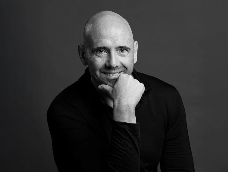 Black and white portrait of a bald man smiling, resting his chin on his hand, wearing a dark shirt.