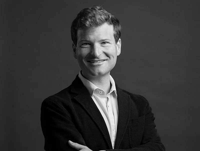 Black and white portrait of a smiling man in a white shirt and dark blazer against a plain background.
