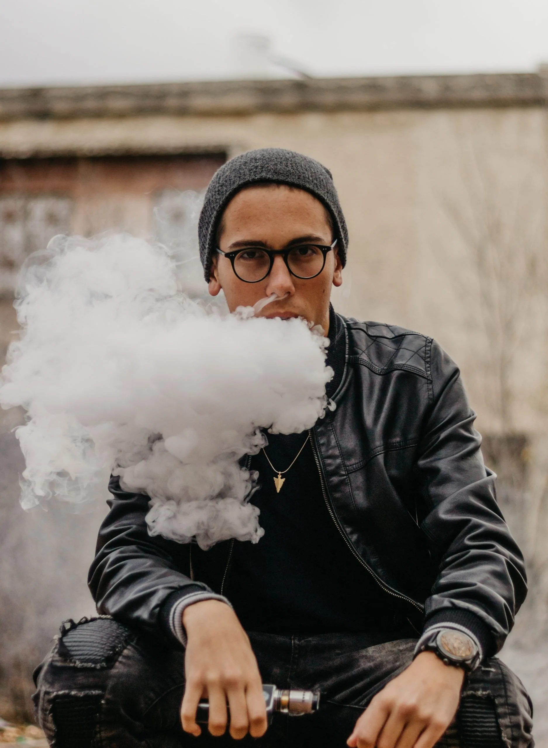 Young man wearing glasses, a gray beanie, and a black leather jacket sitting outdoors. He is holding a vaping device in his right hand, and vapor is emanating from his mouth and the device.