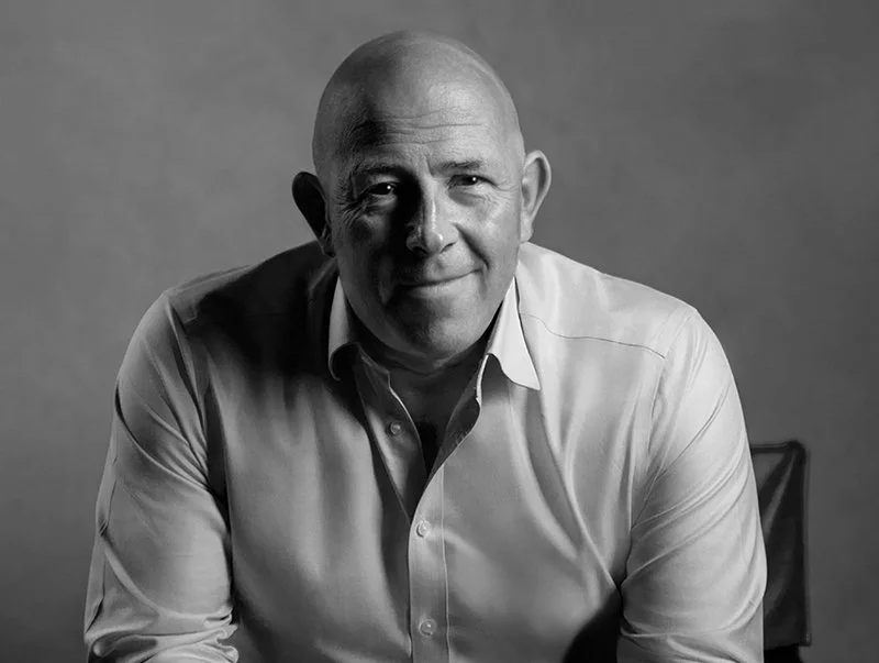 Black-and-white portrait of a middle-aged man with a bald head, wearing a light-colored button-up shirt, sitting on a chair and smiling gently against a plain background.