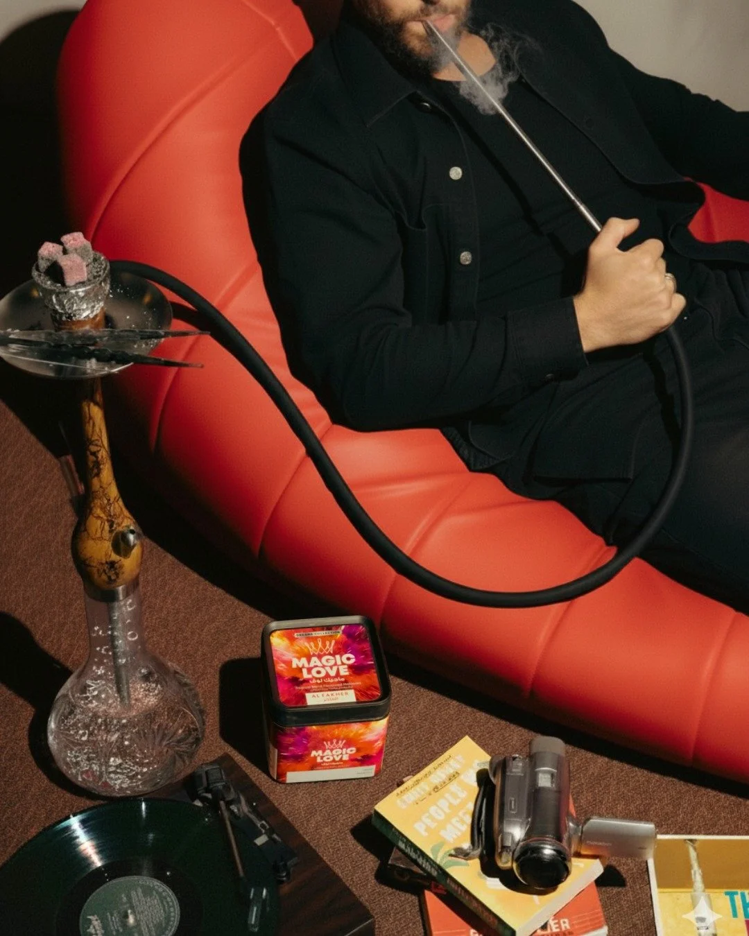 A man is sitting on a red couch, smoking a hookah with a glass base and a long hose. There is another hookah on a table in front of him, along with a box labeled 'Magic Love,' a camera, a vinyl record, and books.