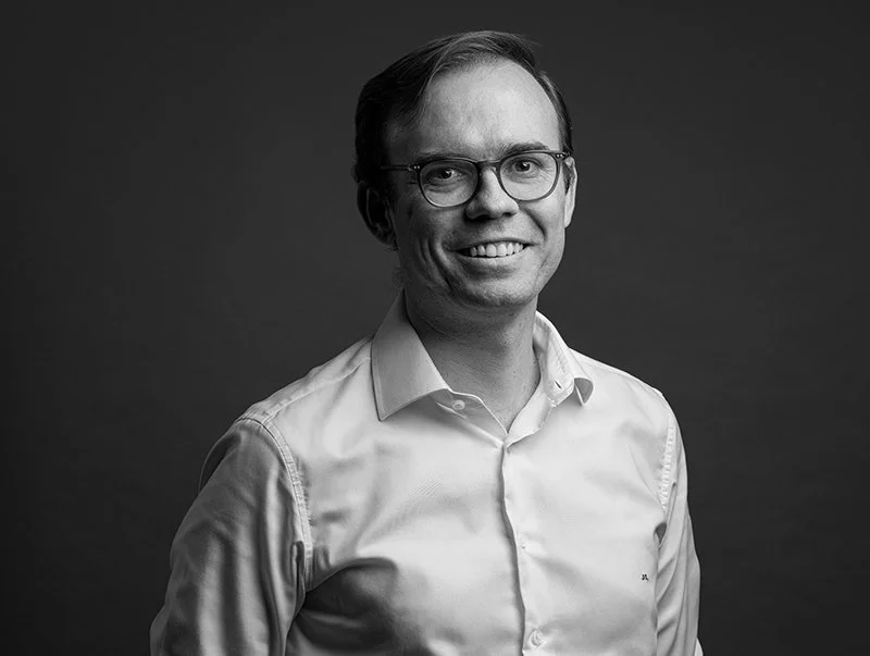 Black and white portrait of a man with glasses, smiling, wearing a collared shirt against a dark background.
