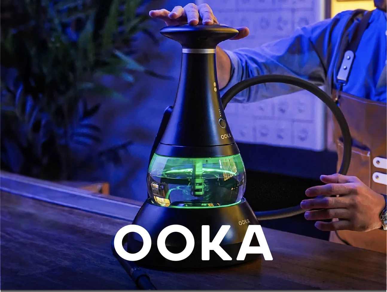 A person using a hookah with a colorful glass base, illuminated with green and blue lights, on a wooden table.