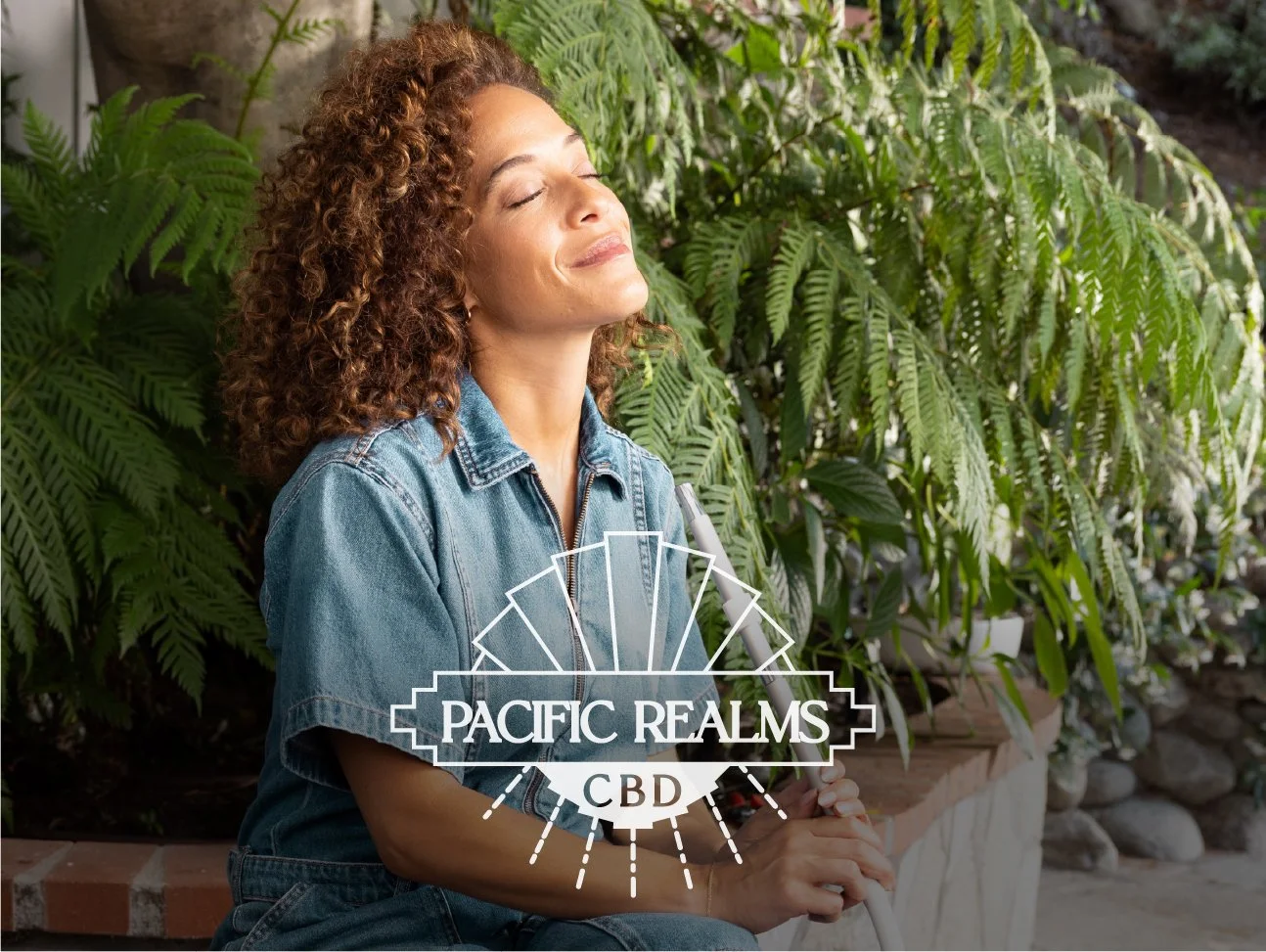 Woman with curly hair wearing a denim shirt, sitting outdoors with green plants behind her, smiling with eyes closed, holding a pen. Overlaid logo says 'Pacific Realms CBD'.