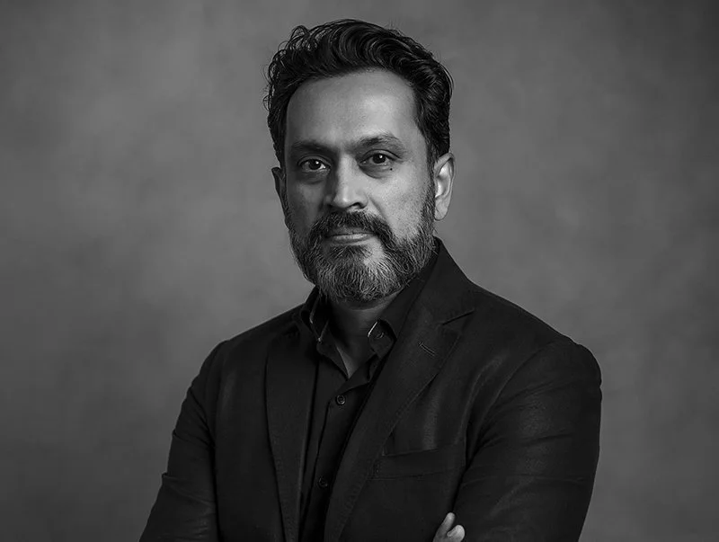 Black and white portrait of a man with dark hair and a beard, wearing a dark blazer and shirt, standing against a neutral background.