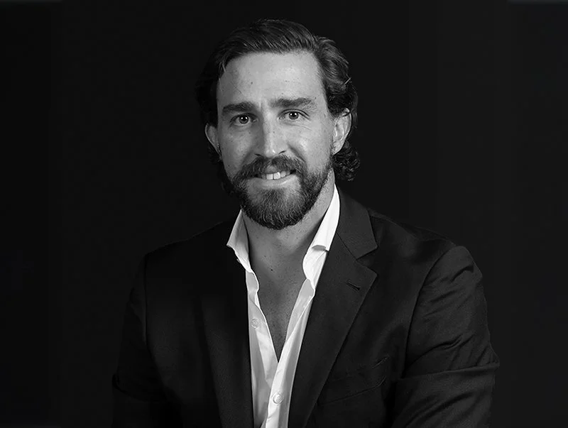 Black and white portrait of a man with dark hair and a beard, wearing a dark suit jacket and white shirt, smiling against a dark background.