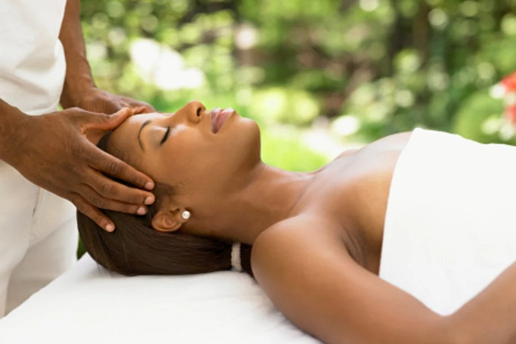 Woman receiving a head massage outdoors, lying on a massage table with greenery in the background.