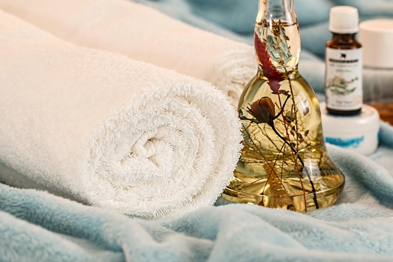 A rolled white towel, a glass bottle with dried flowers inside, and various small bottles of oil or lotion on a soft surface.