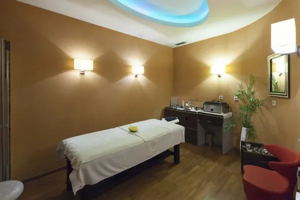 A massage therapy room with a massage table covered in white sheet, a small yellow flower on it, warm beige walls, three wall sconces providing soft lighting, a potted plant, a mirror, and a small table with massage supplies and a chair.