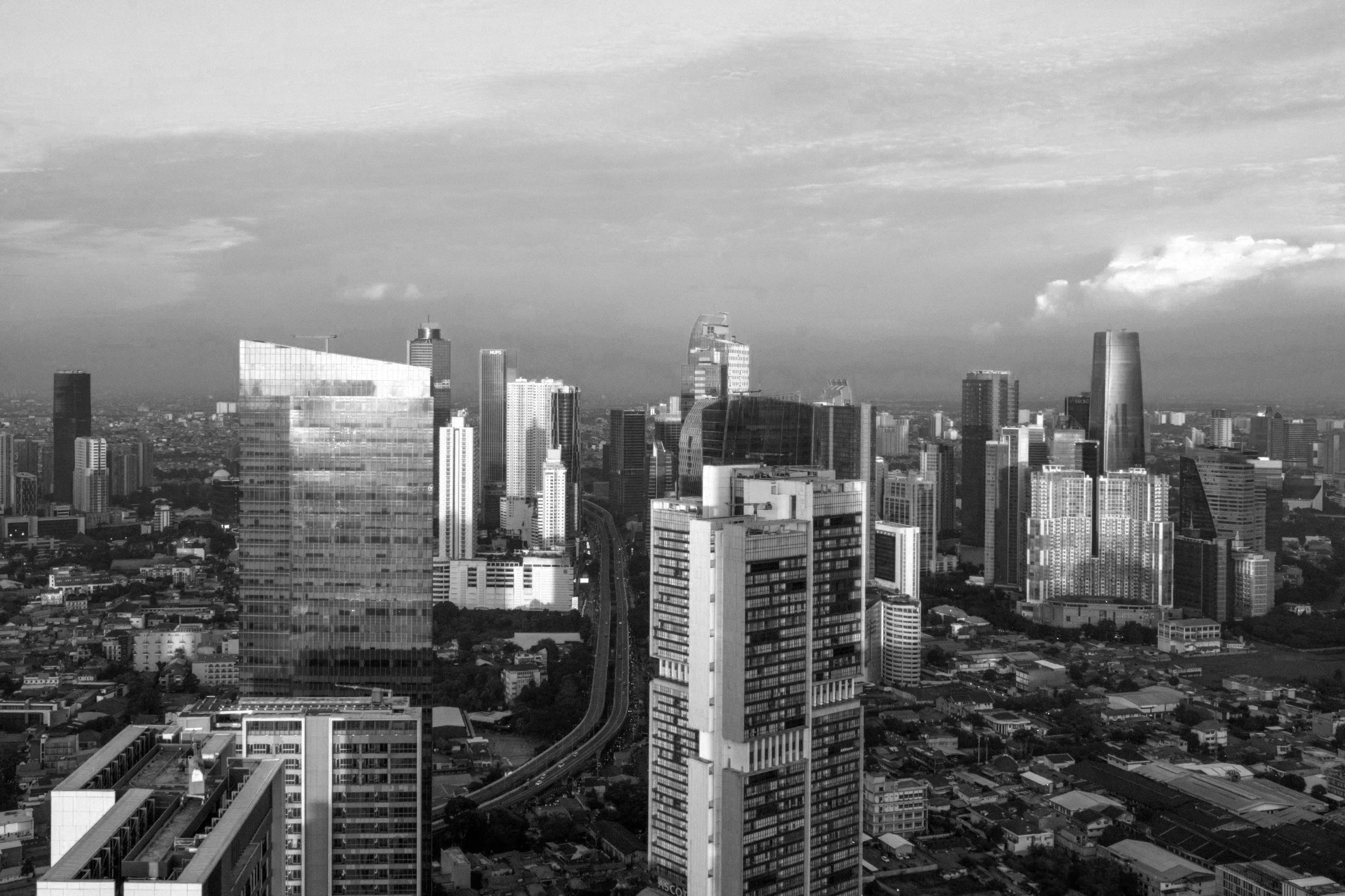 Black and white aerial view of city skyline with numerous tall skyscrapers and buildings, some with reflective glass facades, under a cloudy sky.