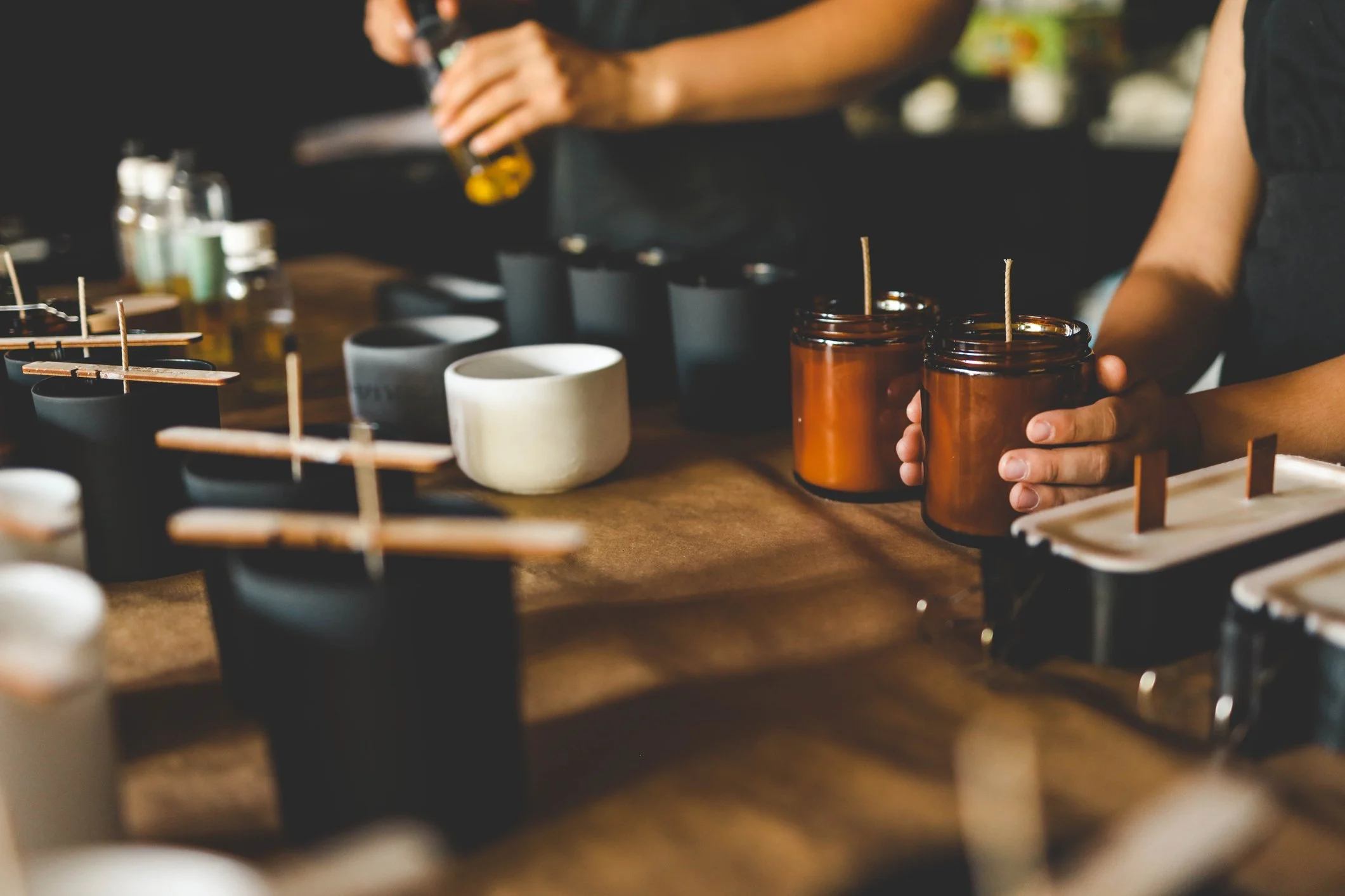 People holding amber glass candle jars with unlit wicks on a wooden table, with various black, white, and gray candles or containers nearby.