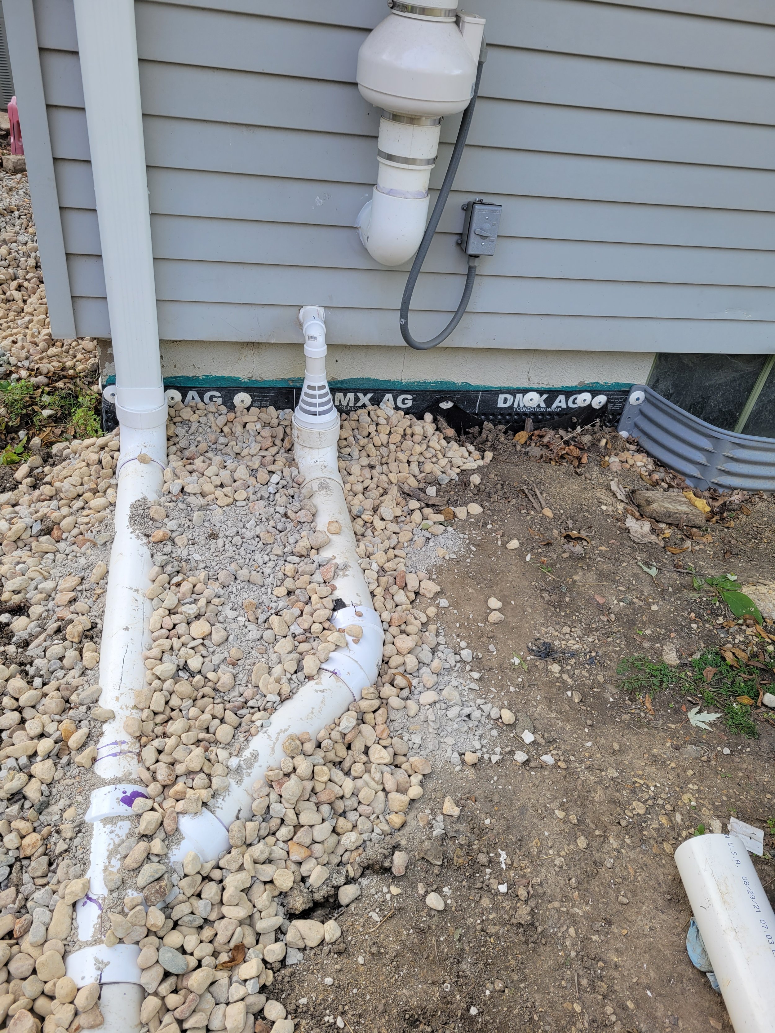 Outdoor view of building foundation with plumbing pipes, gravel, and black foundation drainage wrap