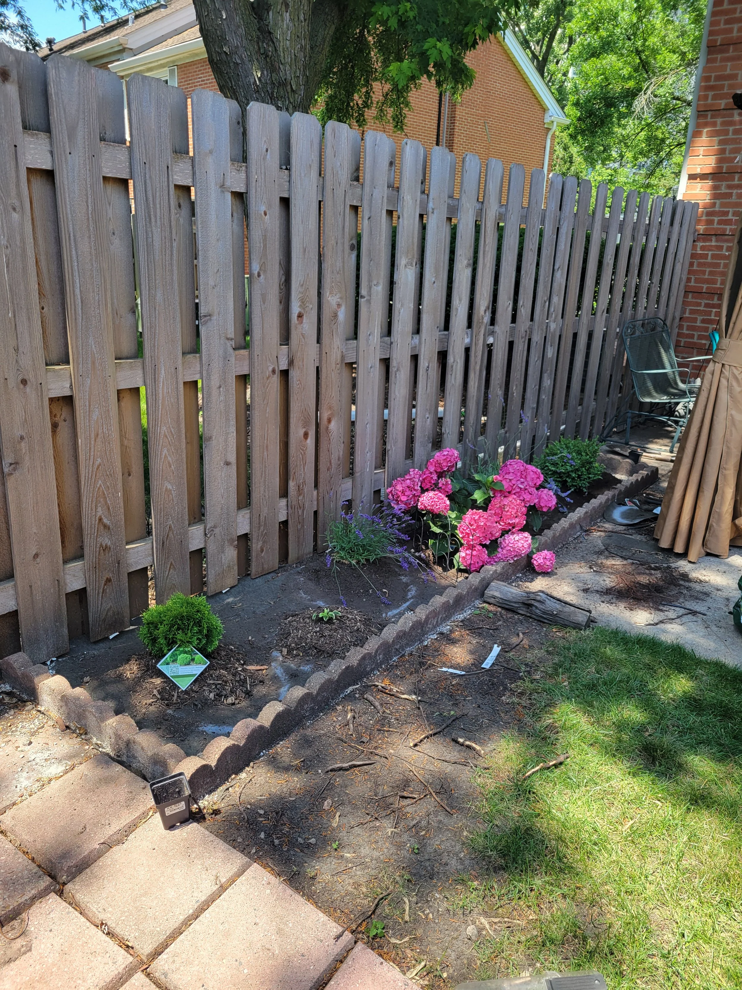 A small garden bed with pink hydrangeas, purple flowers, and some small greenery, bordered by a brick edging, next to a wooden fence. There is a bench and some patio furniture on the right side, with a patio stone pathway in the foreground.