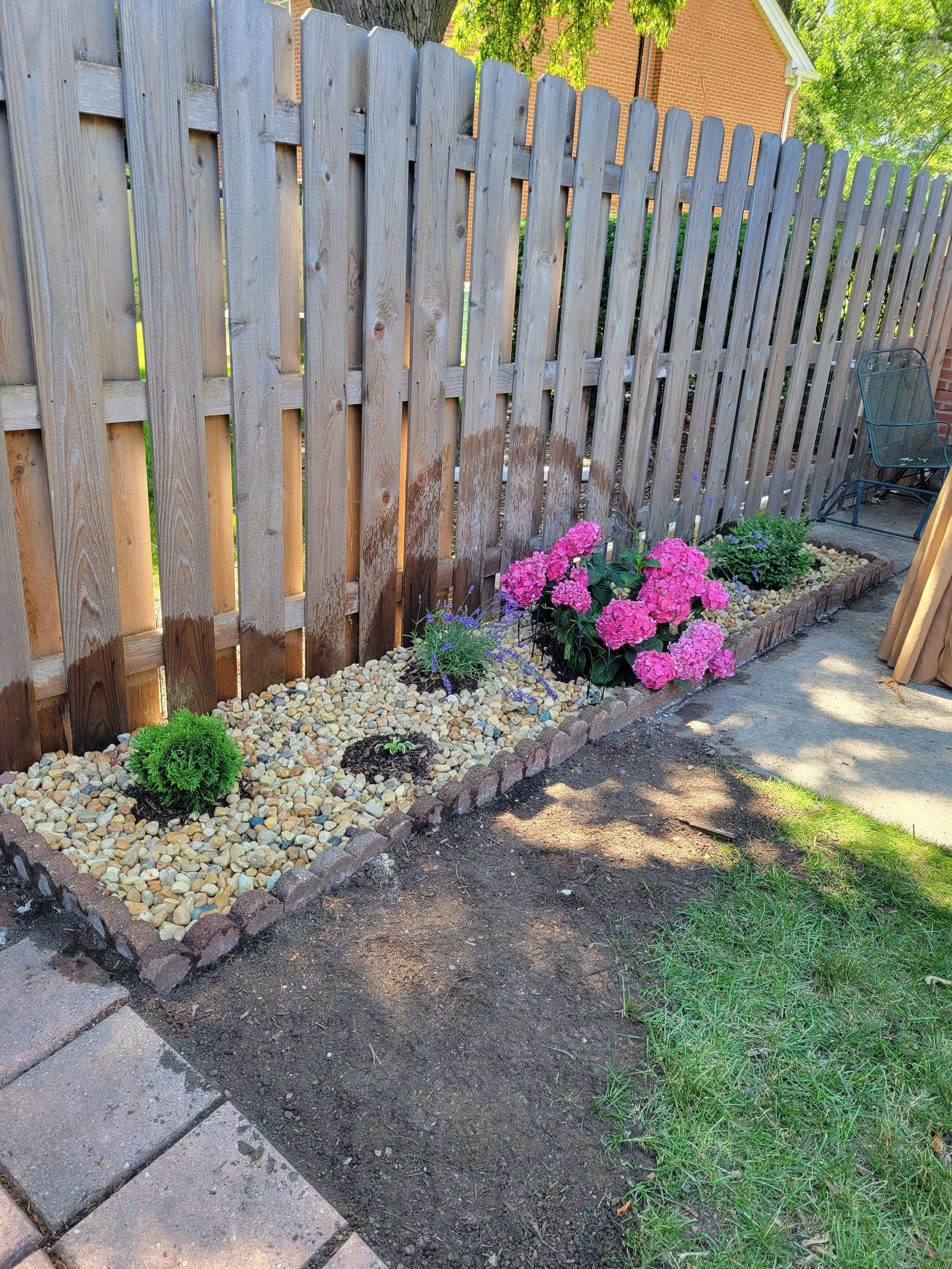 A backyard garden bed with pink hydrangeas, purple flowers, and small evergreens, bordered by bricks and filled with small pebbles, located next to a wooden fence and concrete patio.